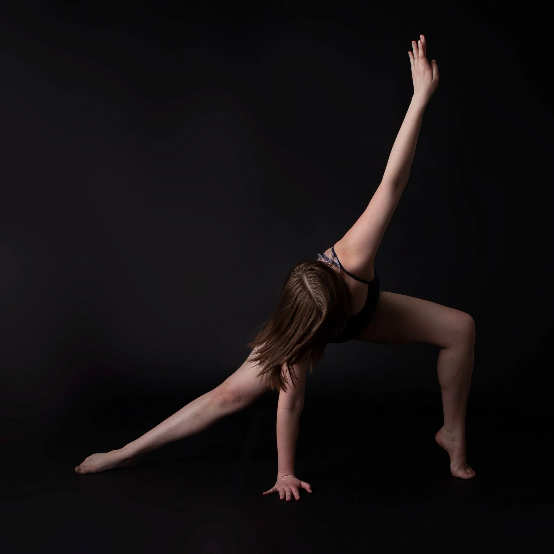 A woman performing a yoga pose, with one hand on the ground, one leg bent with the foot flat, the other leg extended outward, and one arm raised straight up, against a black background.