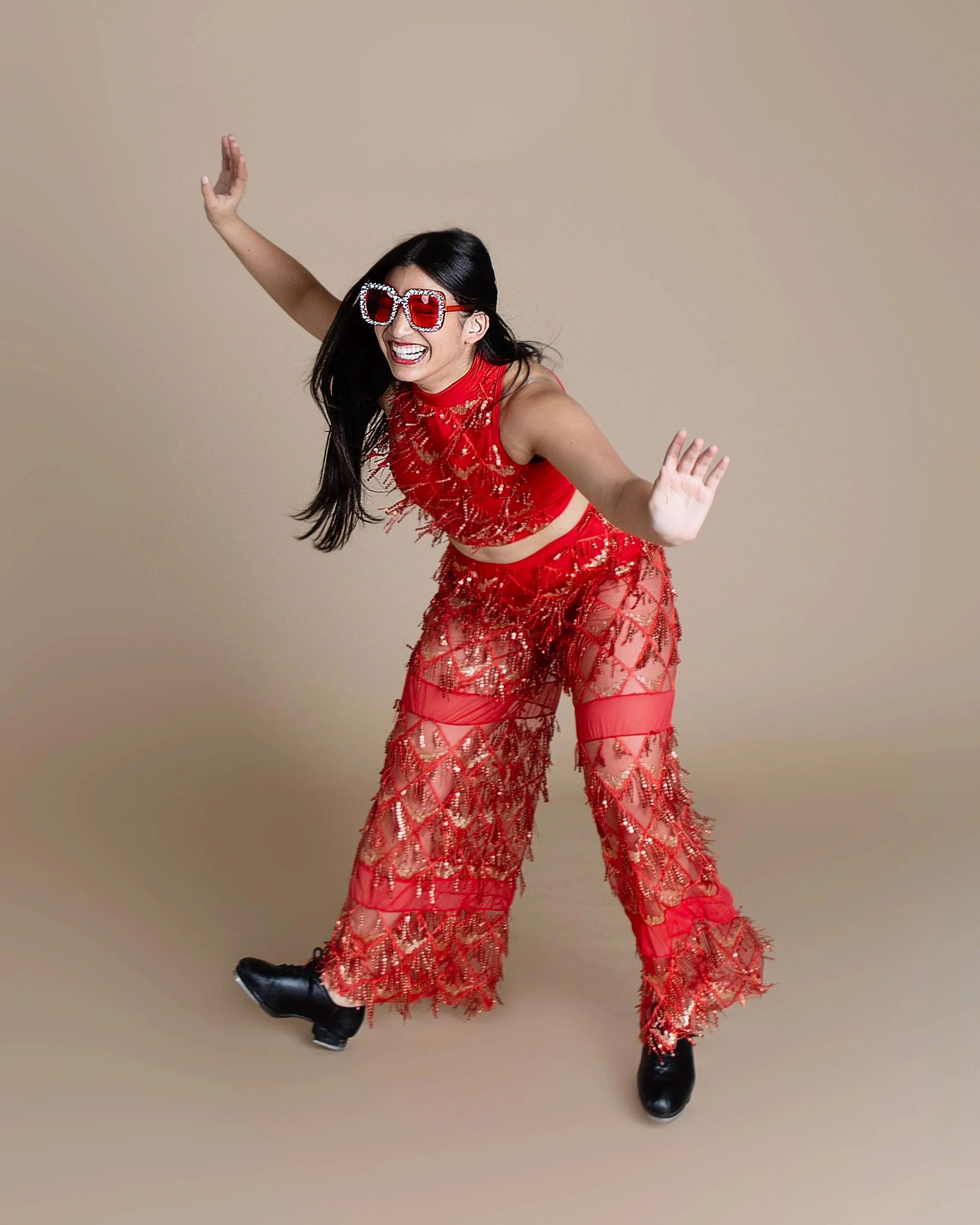 A woman wearing a red, sparkly, and sheer outfit with patterned wide-leg pants, black ice skates, red sunglasses, and smiling playfully against a beige background.