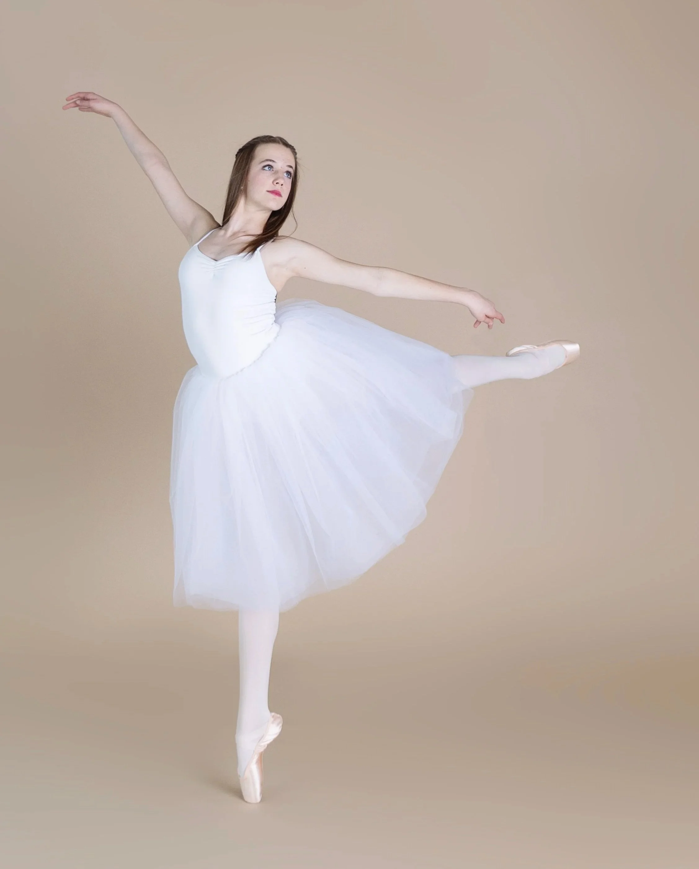 Ballet dancer in a white tutu performing a pose on pointe against a beige background.