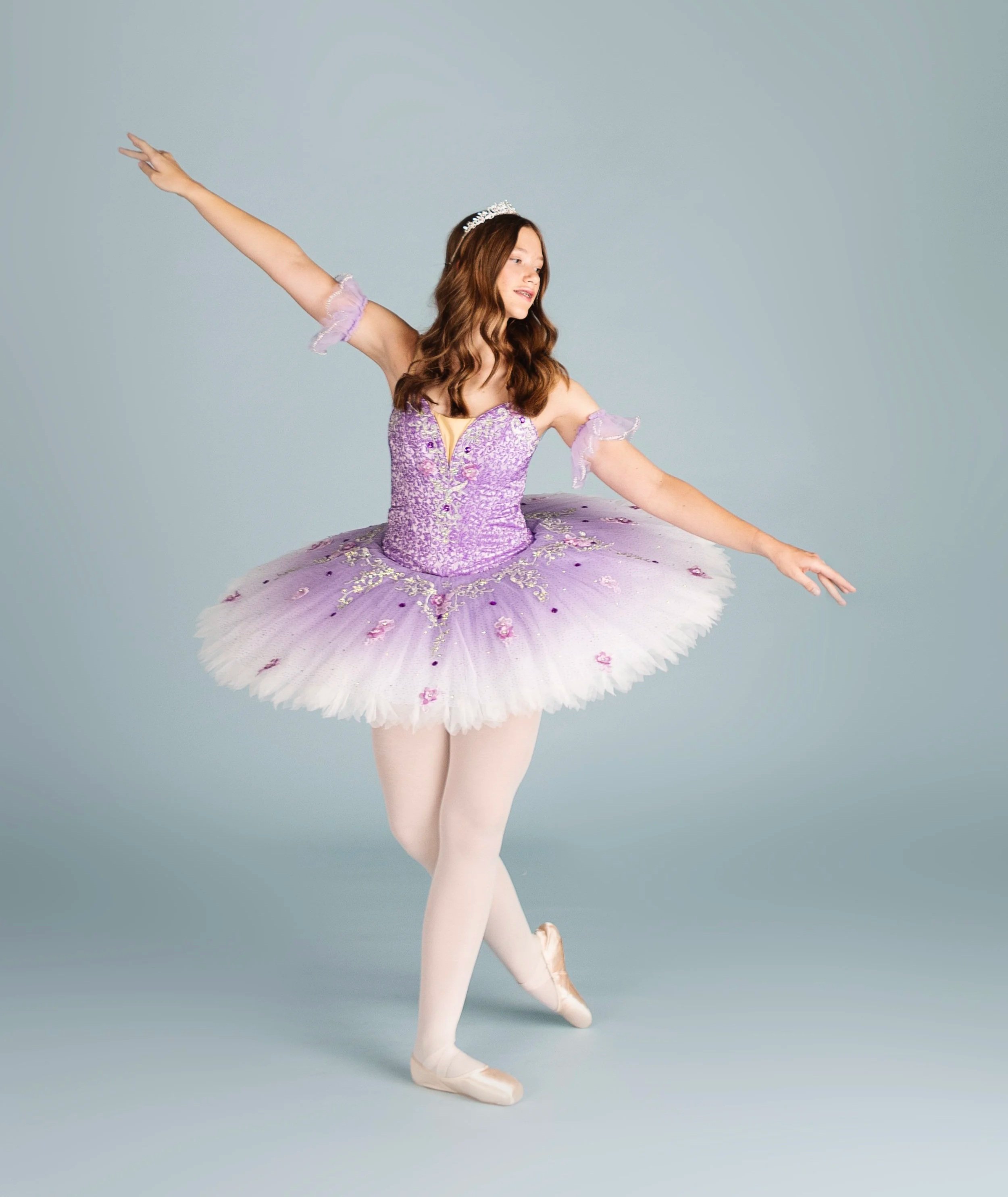 A young woman dressed as a ballerina in a purple tutu with floral embellishments, wearing ballet slippers and a tiara, performing a dance pose against a light gray background.