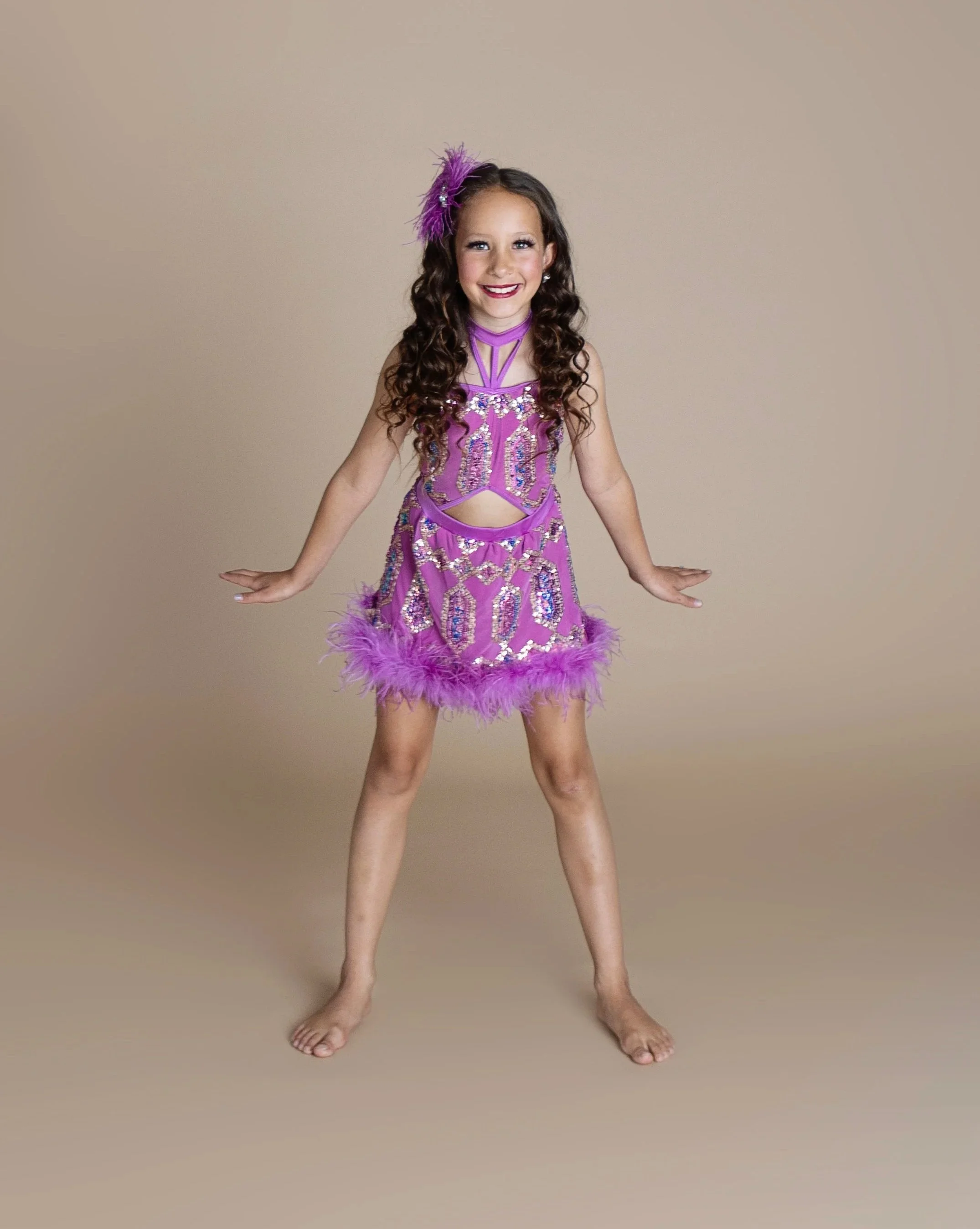 A young girl in a purple, sequined dance costume with feathered trim, smiling and standing barefoot against a plain beige background.