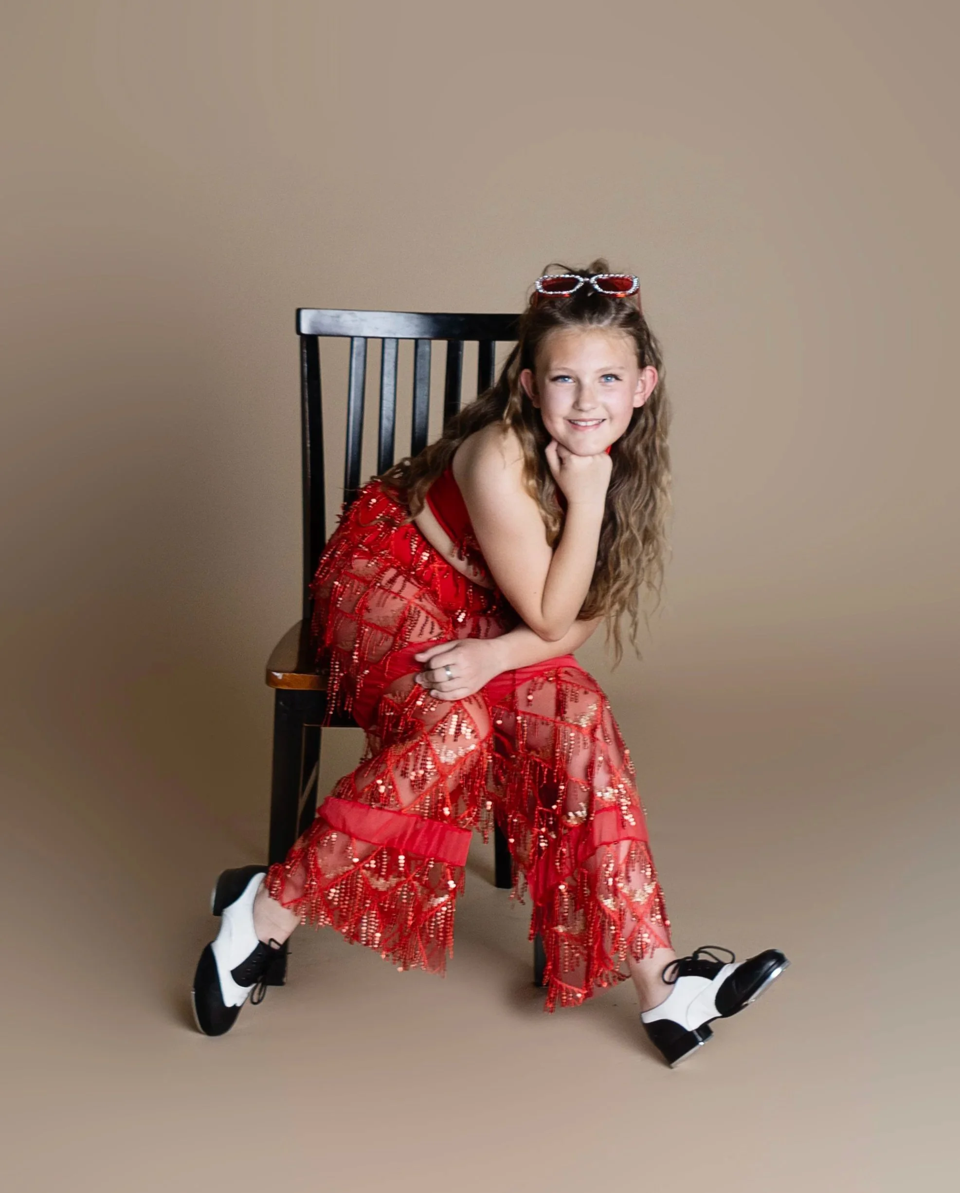 A girl with long curly hair sitting on a black wooden chair, wearing a red semi-transparent sequined dress with pants underneath, white and black dance shoes, and red sunglasses on her head, smiling at the camera against a plain beige background.