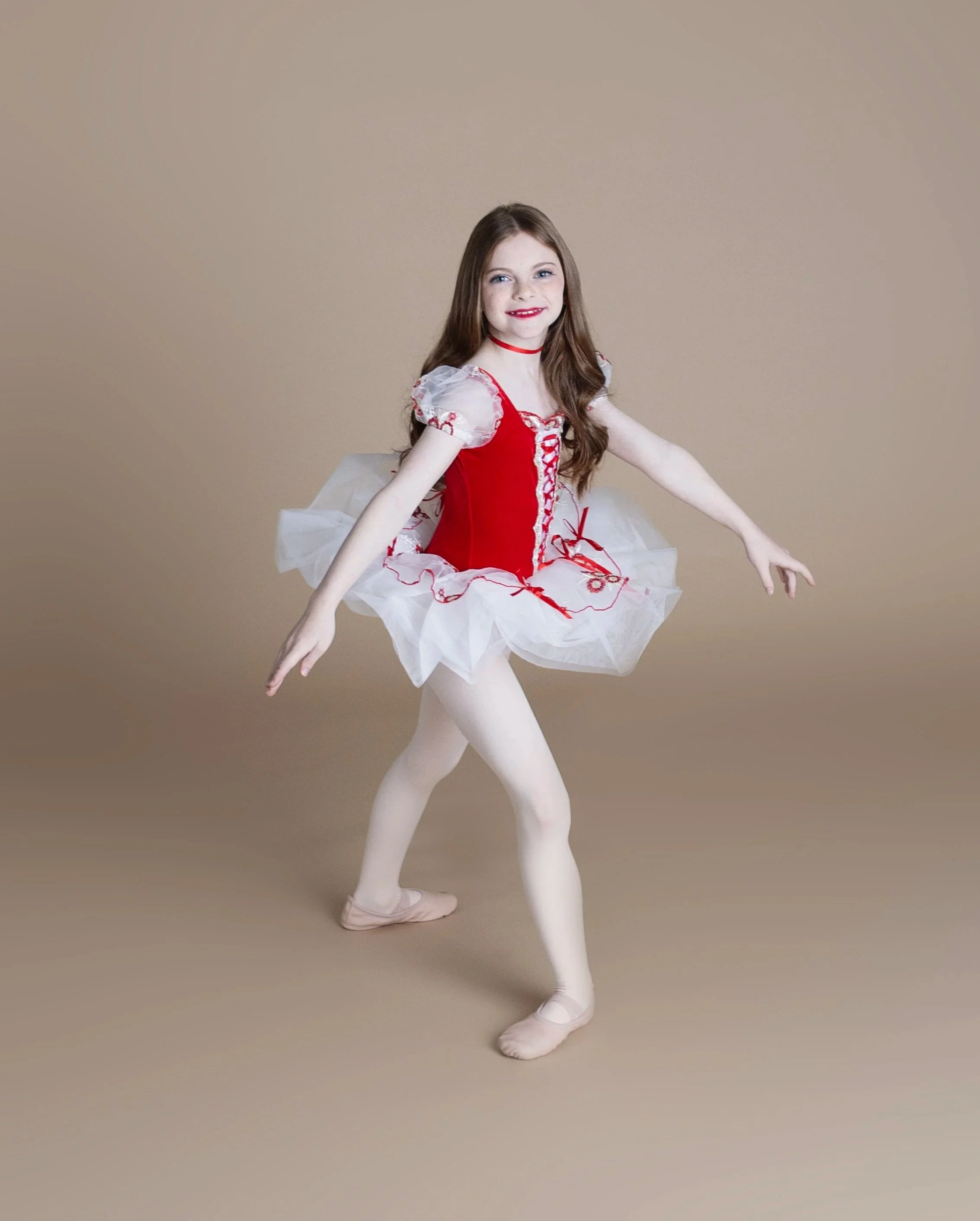 Young girl in a red and white ballet costume, ballet slippers, dancing on plain beige background.