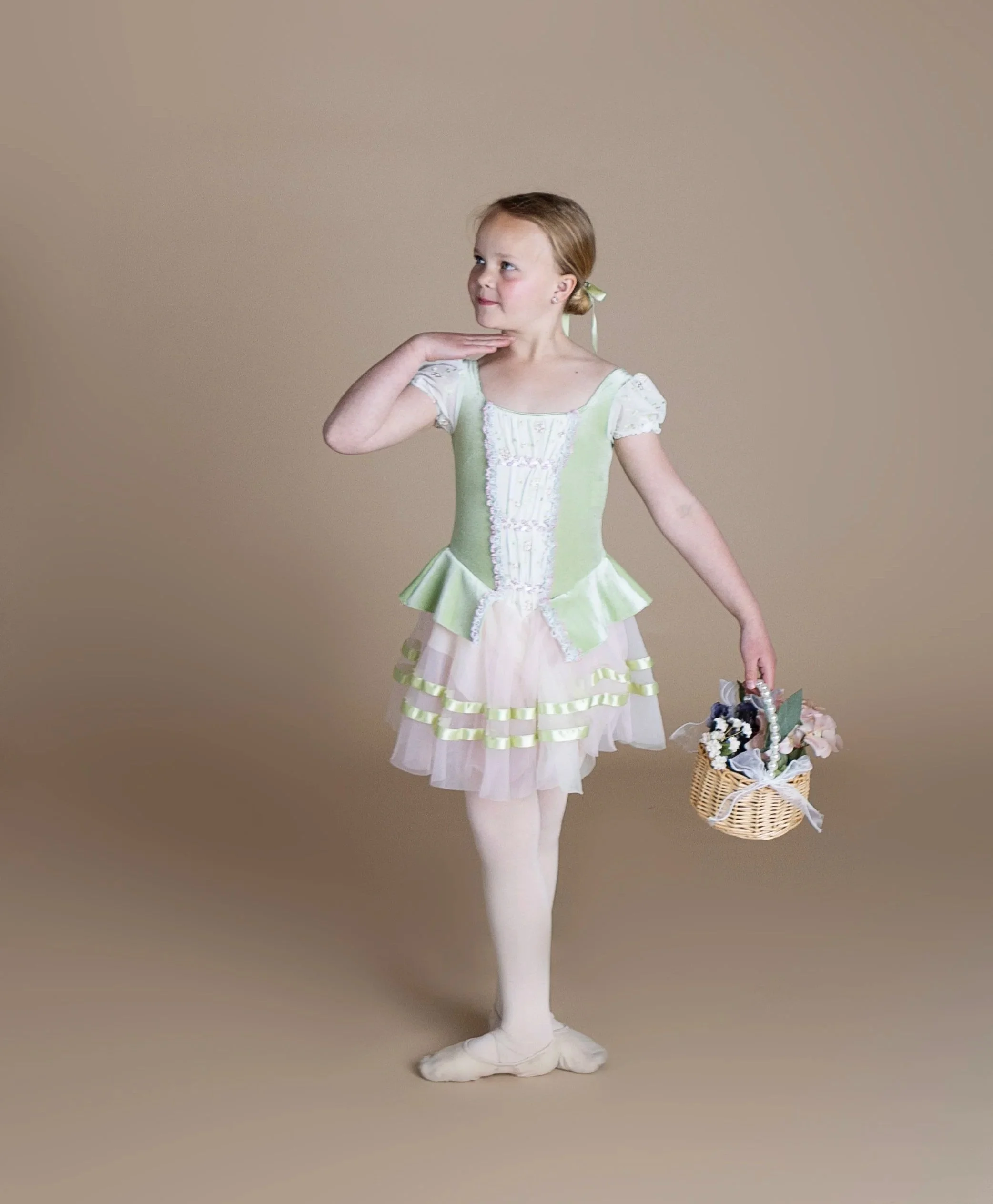 A young girl in a light green and cream dress with ruffles, holding a small basket with flowers, standing on a plain background.