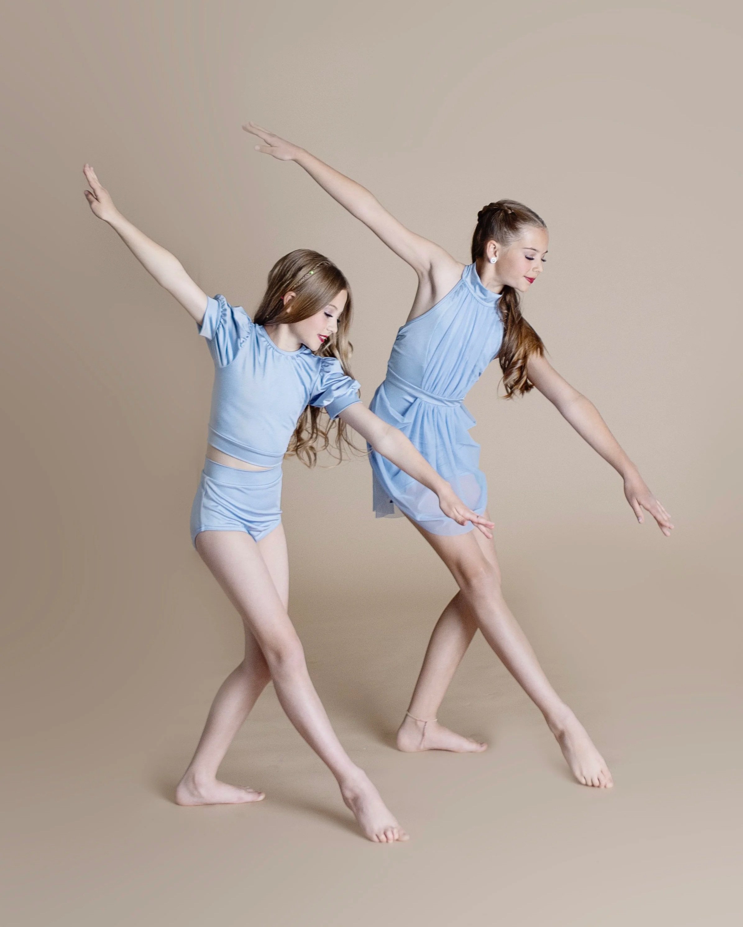 Two young girls dance together in matching light blue dresses, striking a stylized pose against a neutral background.