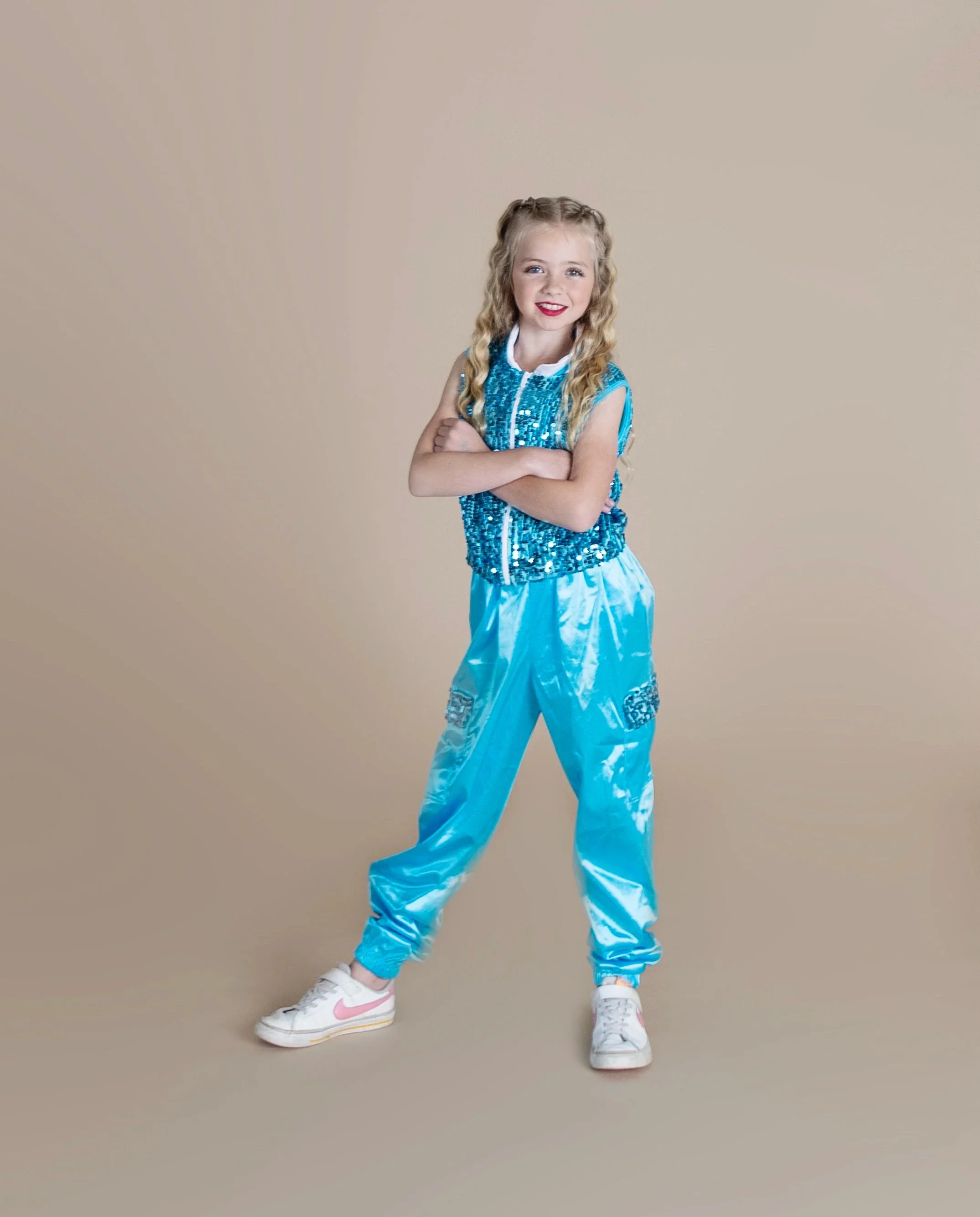 A young girl with long curly blonde hair, wearing a blue sequined top and shiny light blue pants, stands with arms crossed and smiles at the camera.