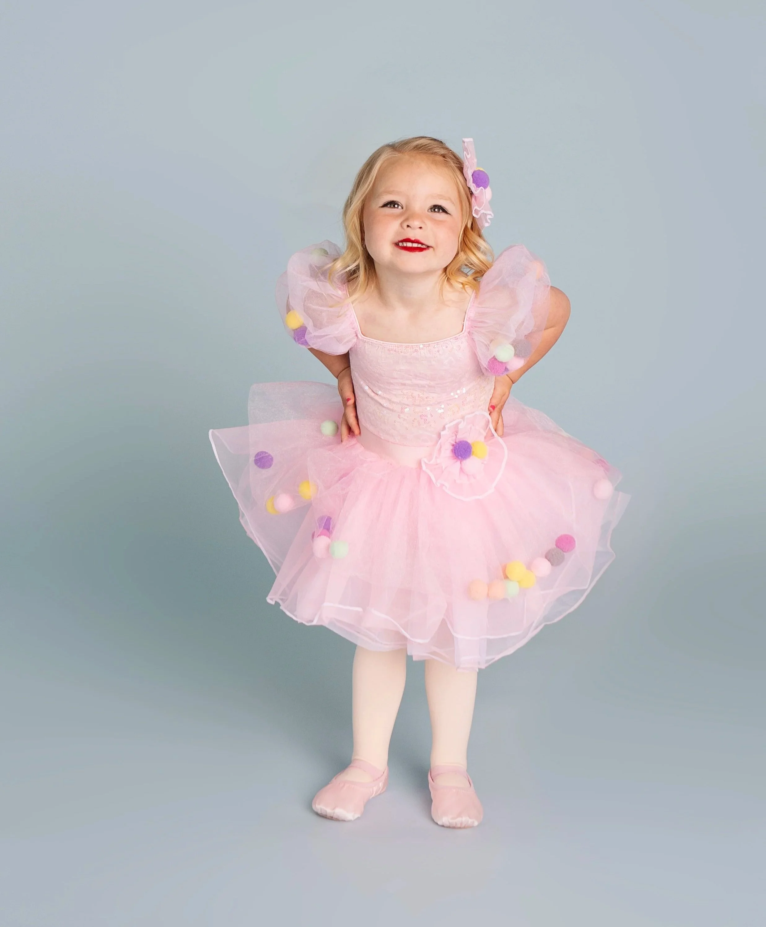 A young girl wearing a pink tutu dress with multicolored pom-poms, standing with hands on her hips, smiling, against a plain grey background.