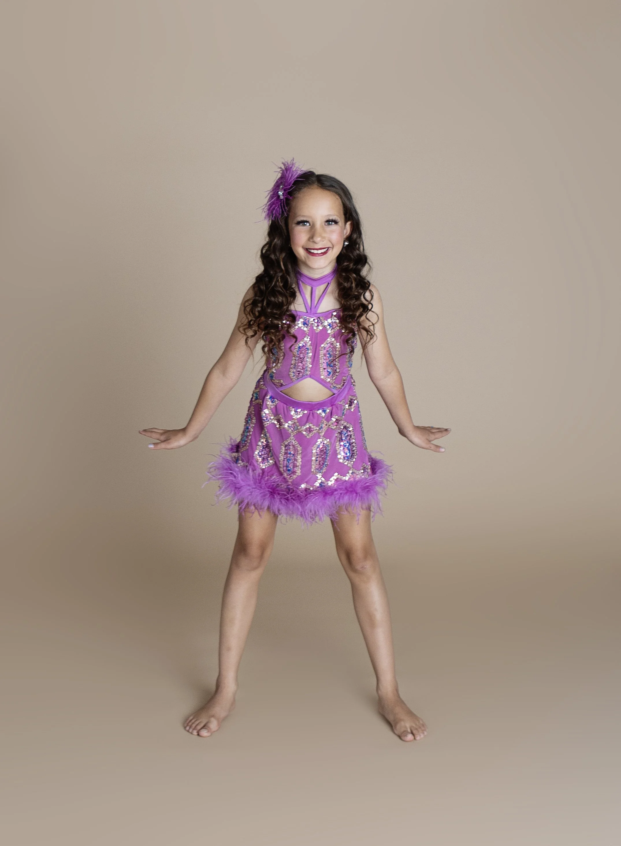 A young girl in a purple, sequined dance costume with feathered trim, smiling and standing barefoot against a plain beige background.