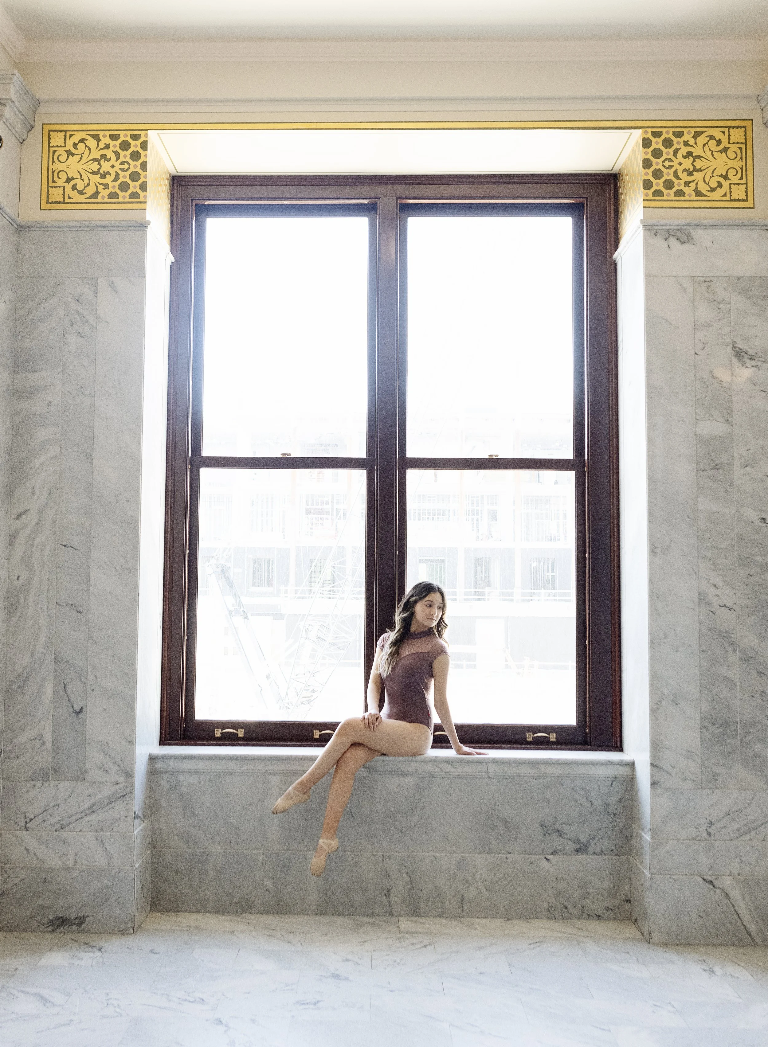 A ballerina in a mauve leotard and beige tights sitting on a wide marble ledge by a large window, with a cityscape visible outside.