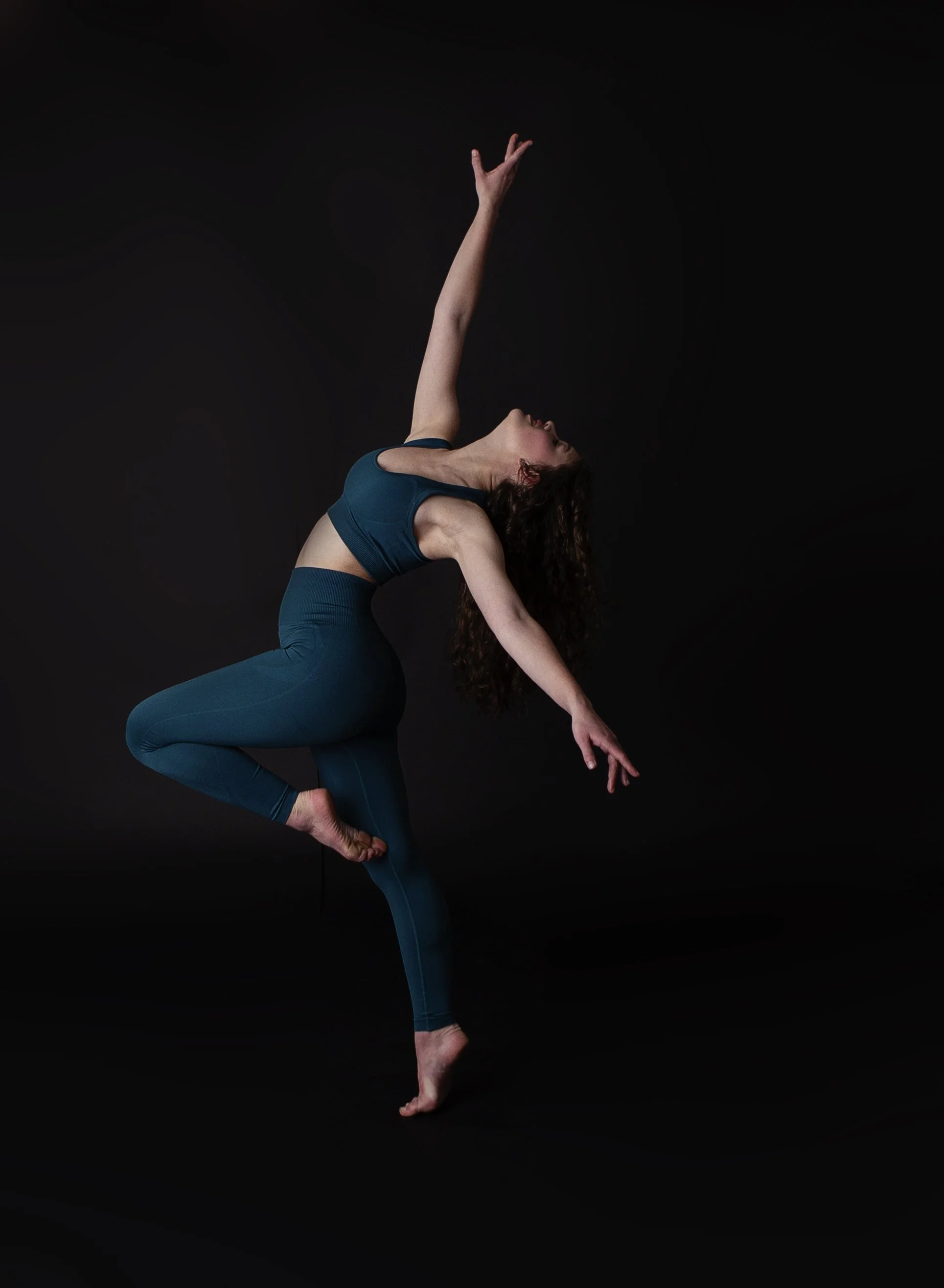 A woman performing a dance or yoga pose against a black background, extending one arm upward and the other downward, with one leg pulled up and the other supporting her balance.