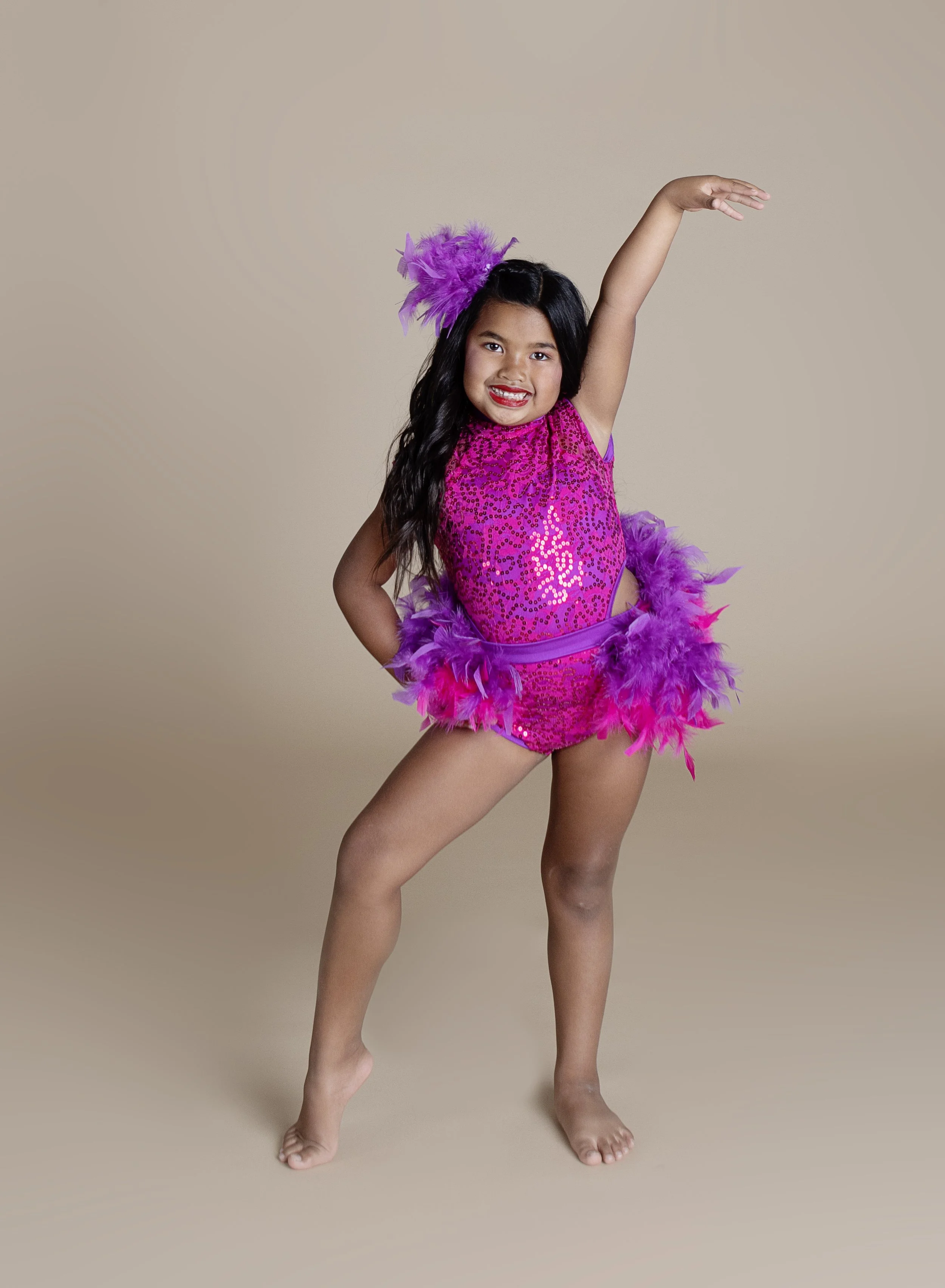 Young girl dressed in bright pink and purple costume with feathers, smiling with one arm raised in a dance pose.