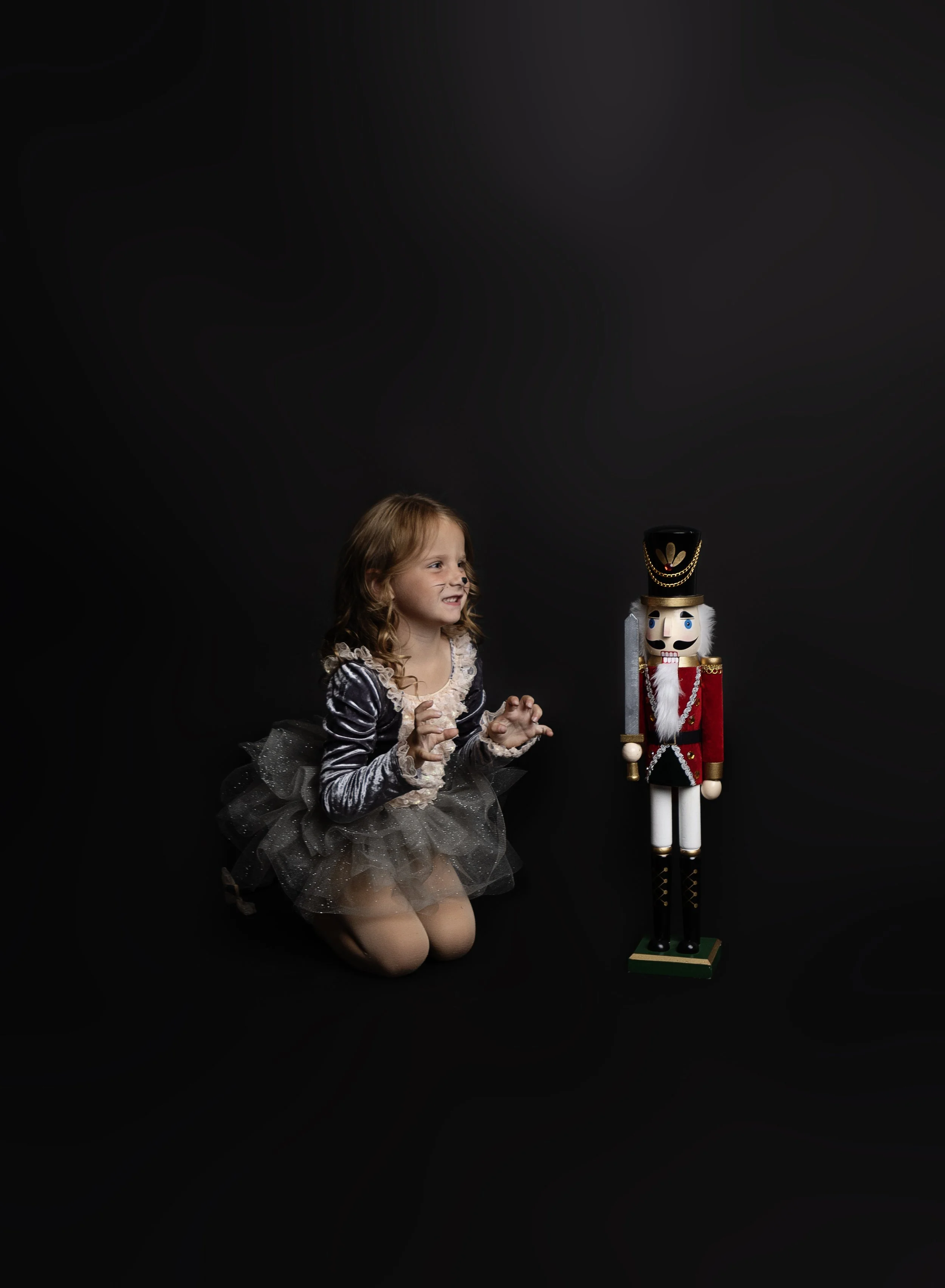 A young girl dressed in a sparkly gray dress with a tutu, kneeling and smiling next to a large nutcracker figure against a black background.