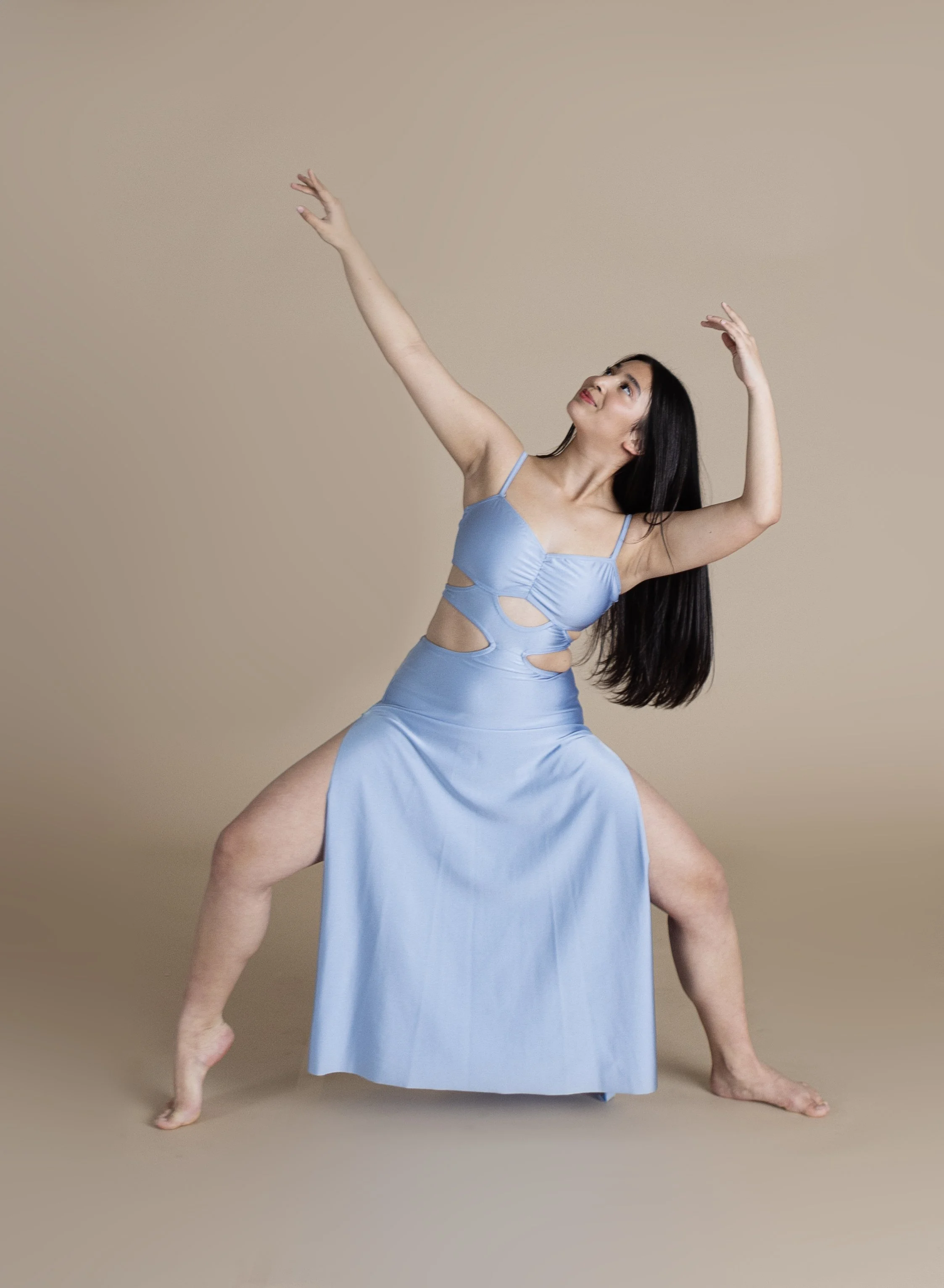 A woman wearing a light blue dress with cut-outs, striking a dance pose with arms raised and body leaning to one side against a beige background.