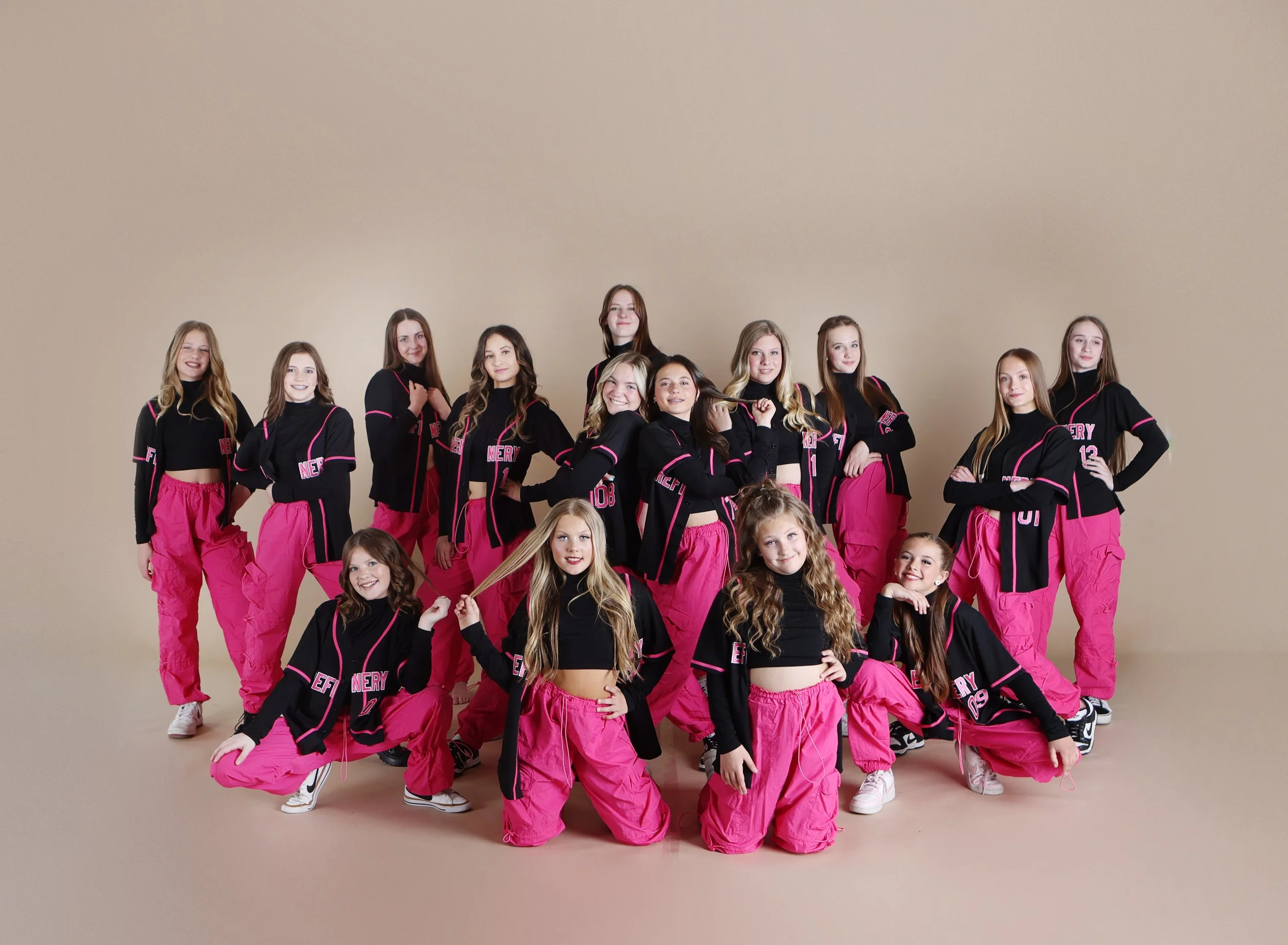 Group of young women in matching dance team outfits with pink pants and black jackets posing together against beige background.