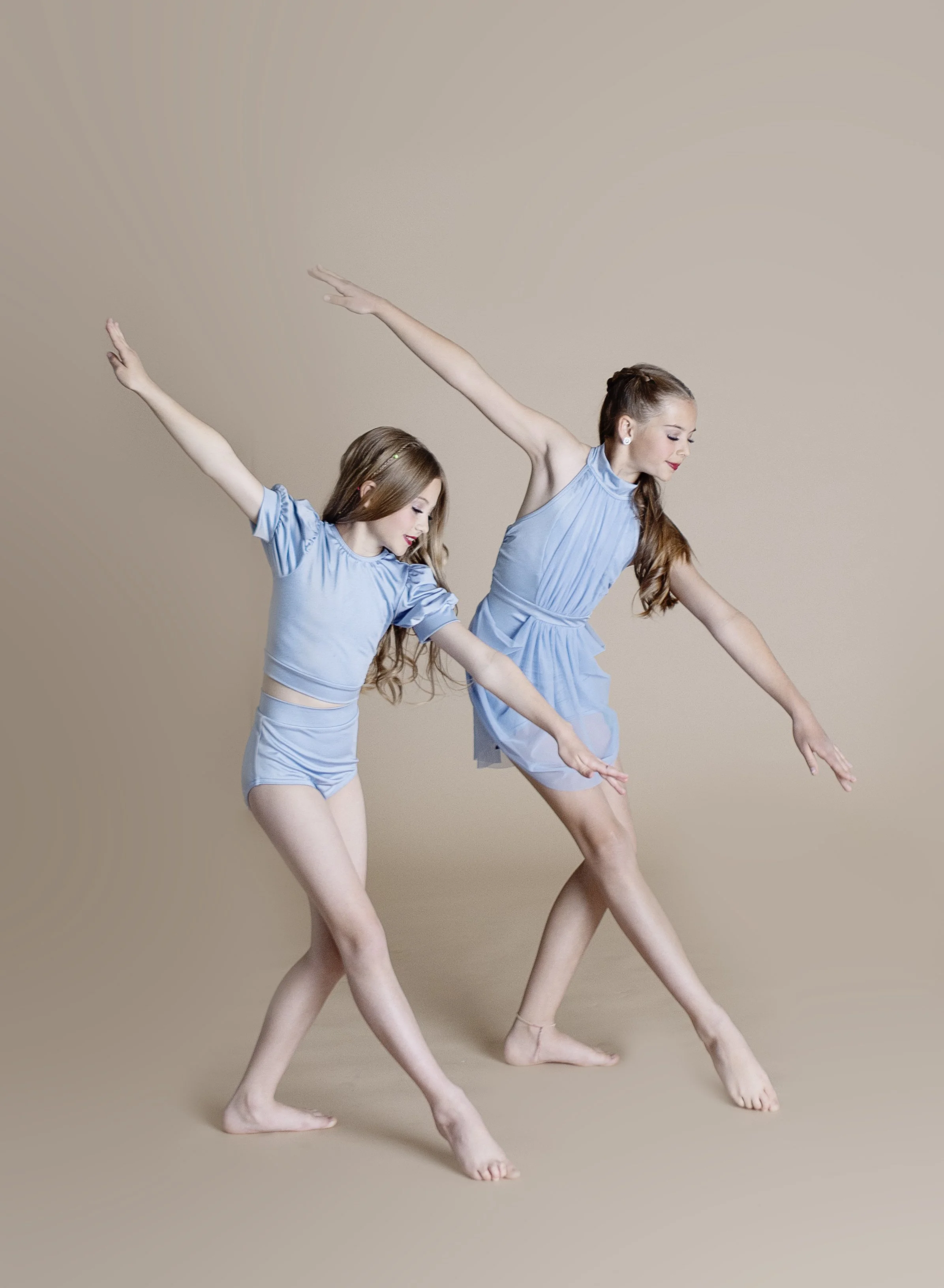 Two young girls dance together in matching light blue dresses, striking a stylized pose against a neutral background.
