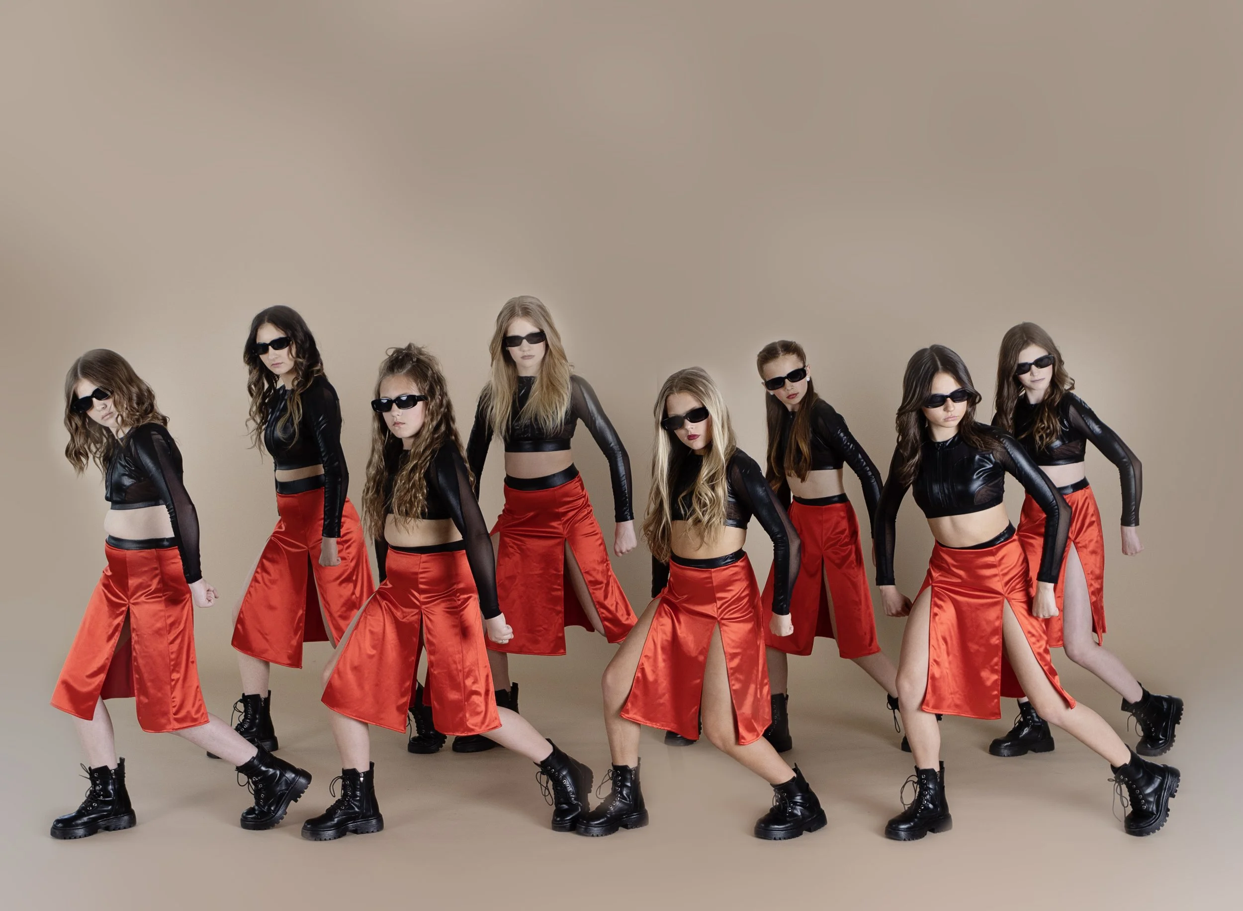 Eight young women dressed in black crop tops, red skirts with slits, black boots, and sunglasses, posing against a beige background.