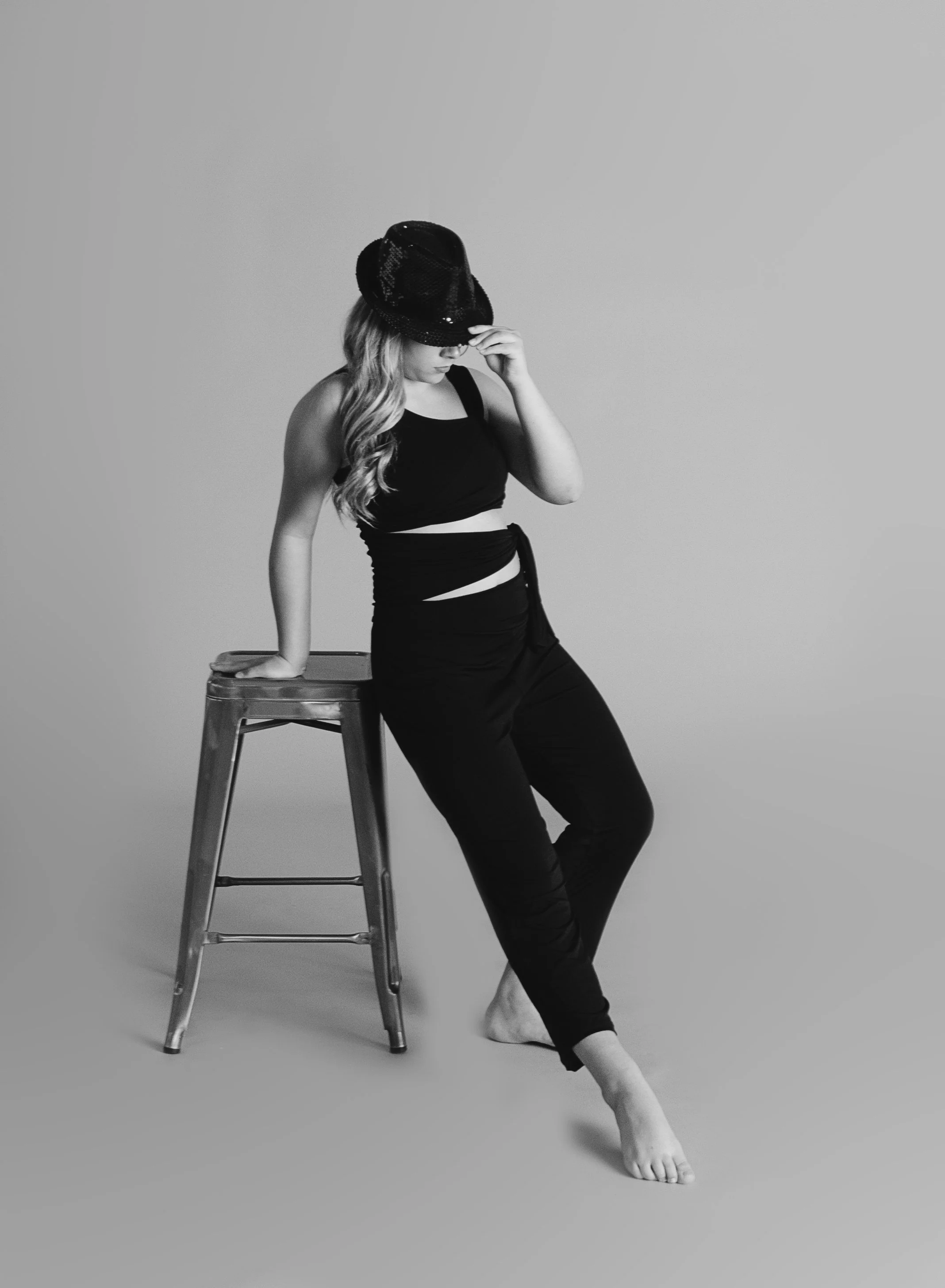 A woman in a black outfit and hat, barefoot, leaning on a wooden stool against a plain background.