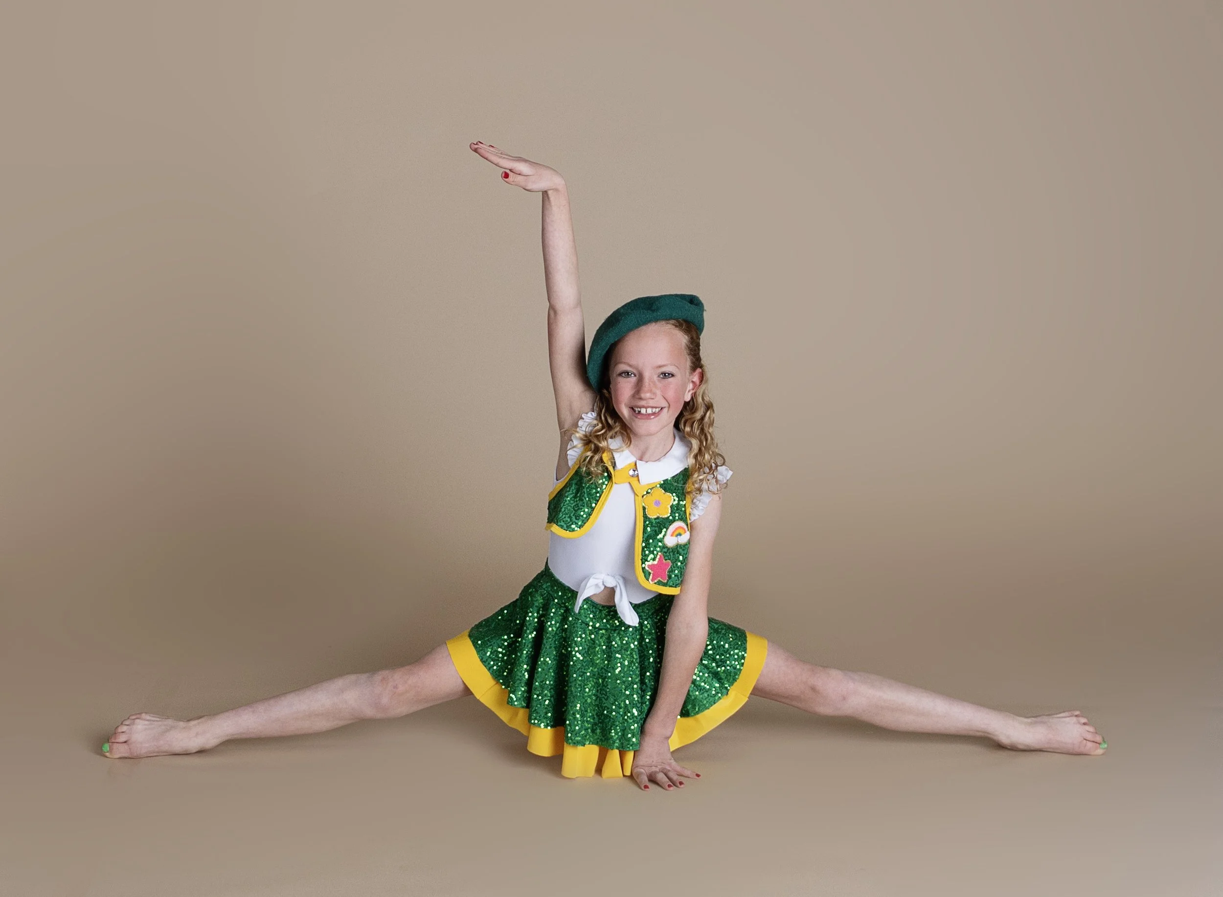 Young girl doing splits on the floor, wearing a green sequined skirt, white top, and a green beret, smiling.