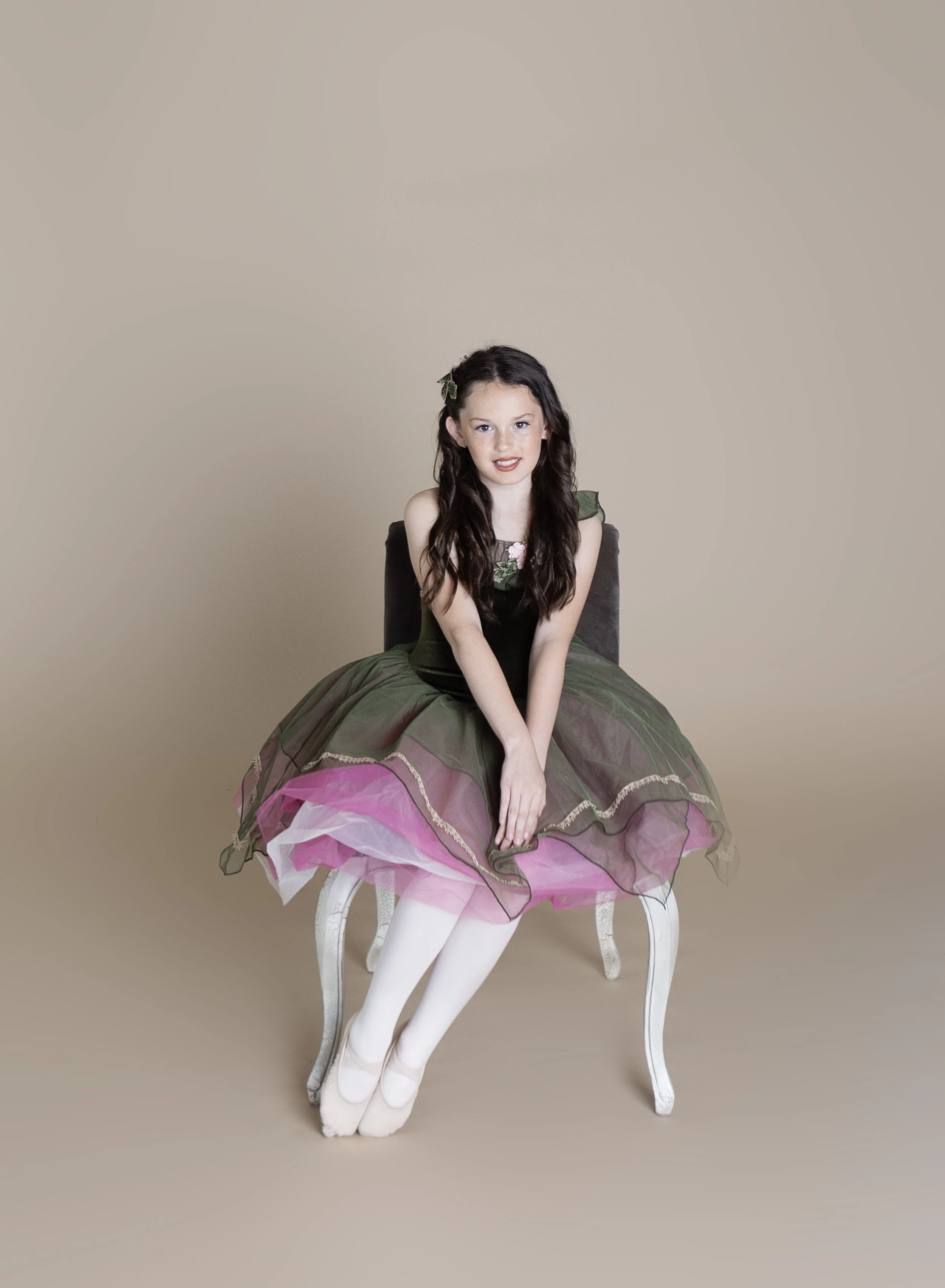 A young girl with long dark hair sitting on a chair, wearing a green and pink tutu dress and white tights, against a plain beige background.