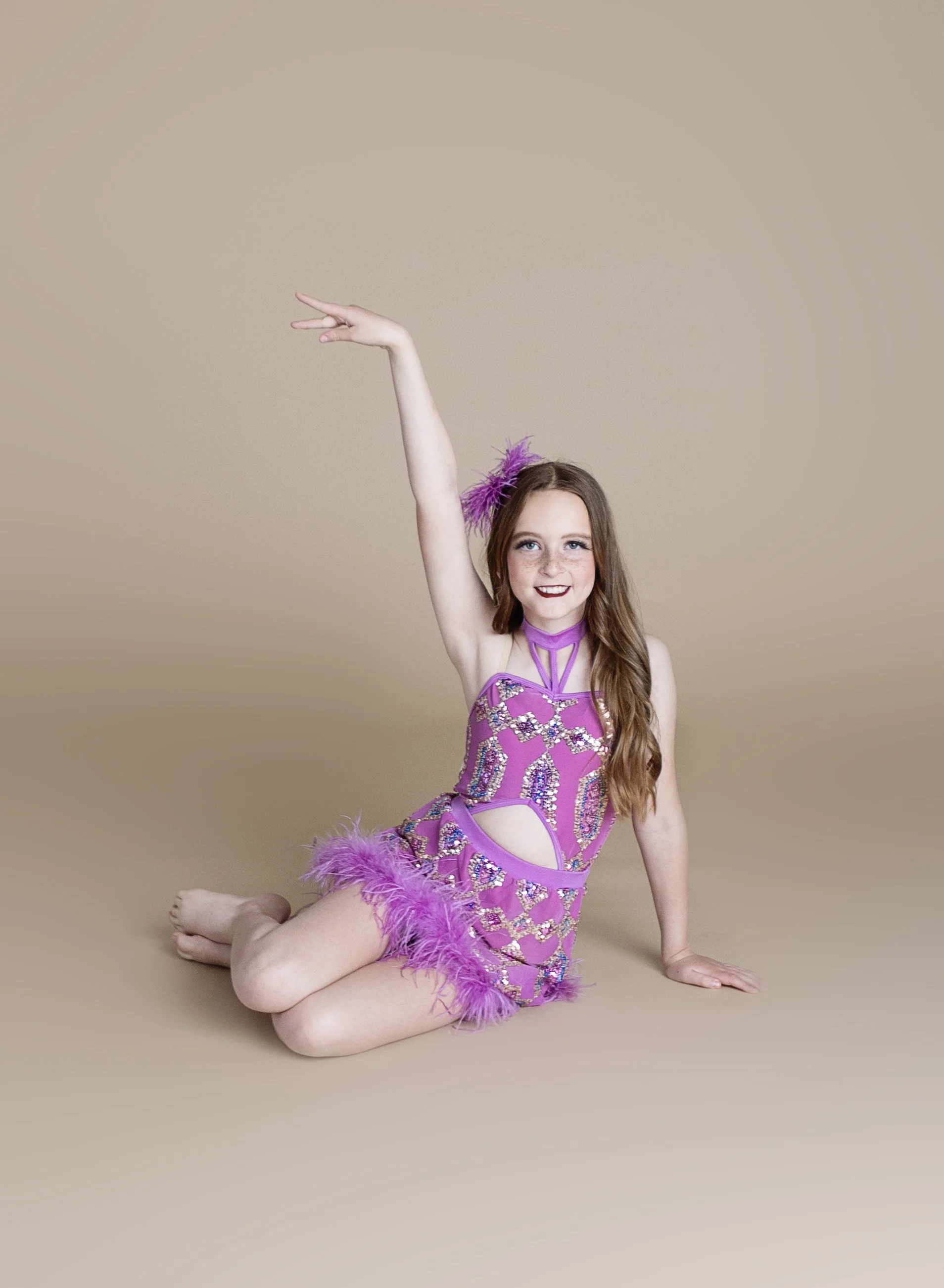 Young girl with long brown hair in a purple sparkly dress with feathers, sitting on beige background, raising her right arm, smiling.