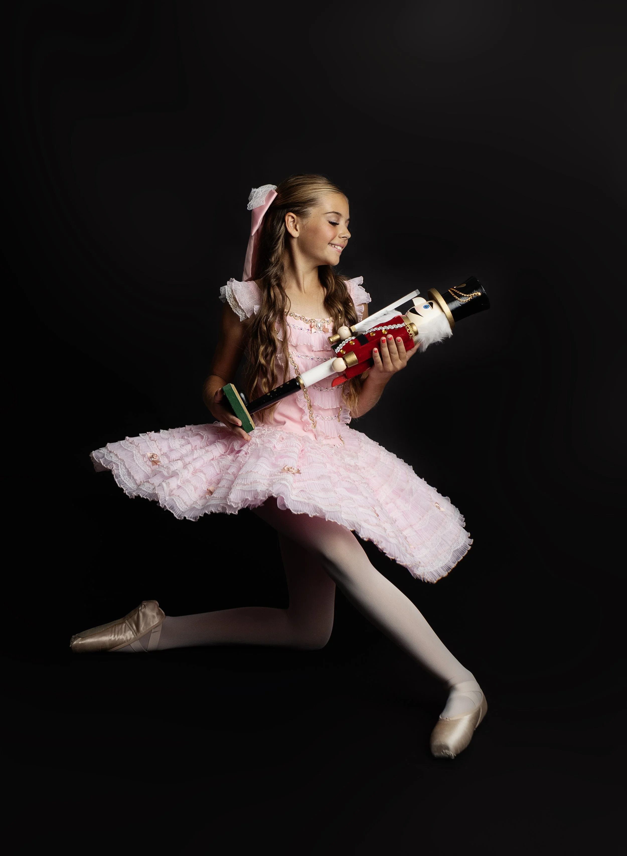 A young girl dressed in a pink ballerina costume holding a nutcracker doll on a black background.