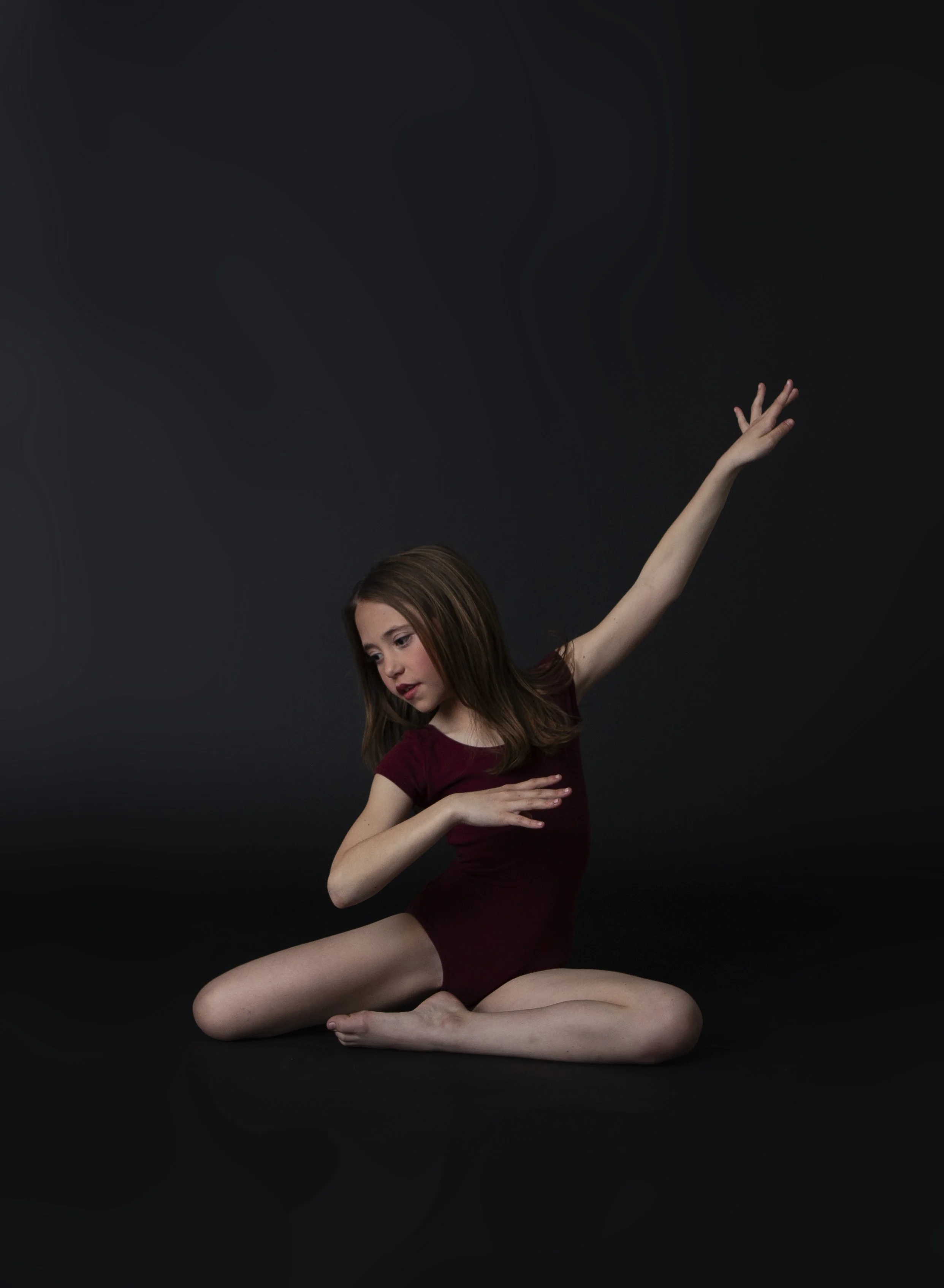 Young girl wearing a maroon leotard sitting on the black floor with her legs crossed, one arm raised above her head, and the other arm across her chest, in a dance pose against a dark background.