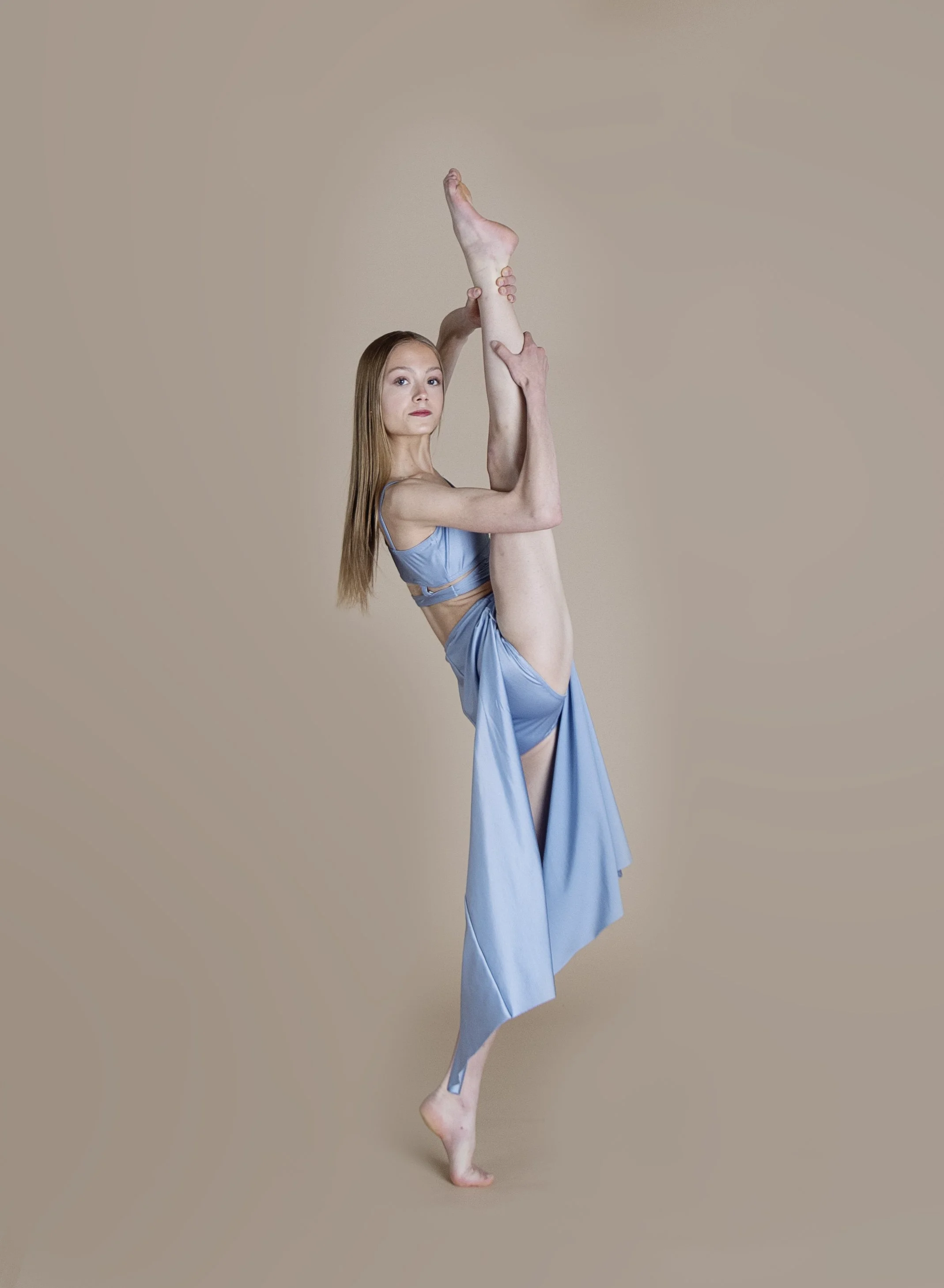 A young woman with long hair in a sky blue dance outfit, performing an expressive dance pose against a beige background.