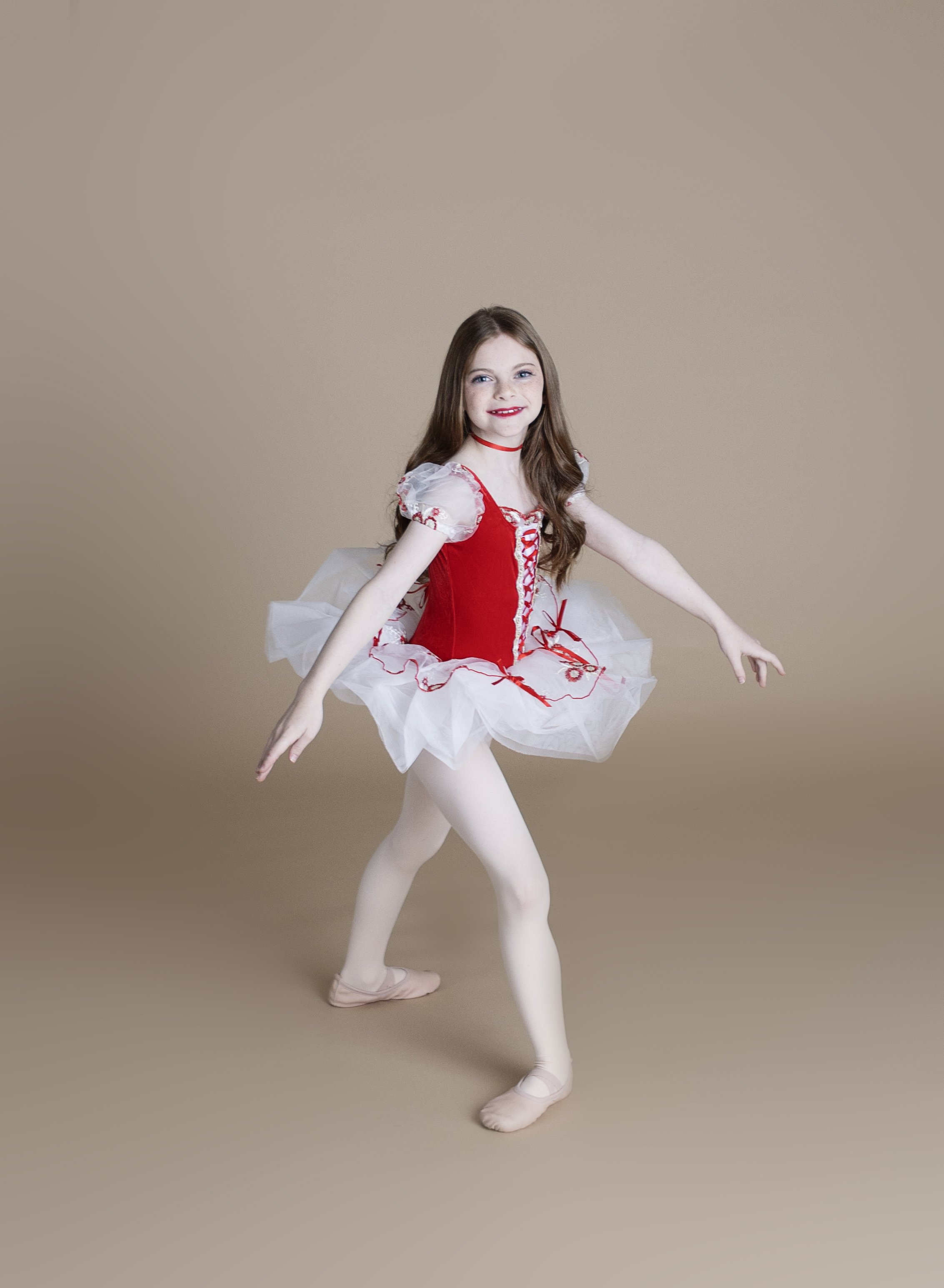 Young girl in a red and white ballet costume, ballet slippers, dancing on plain beige background.