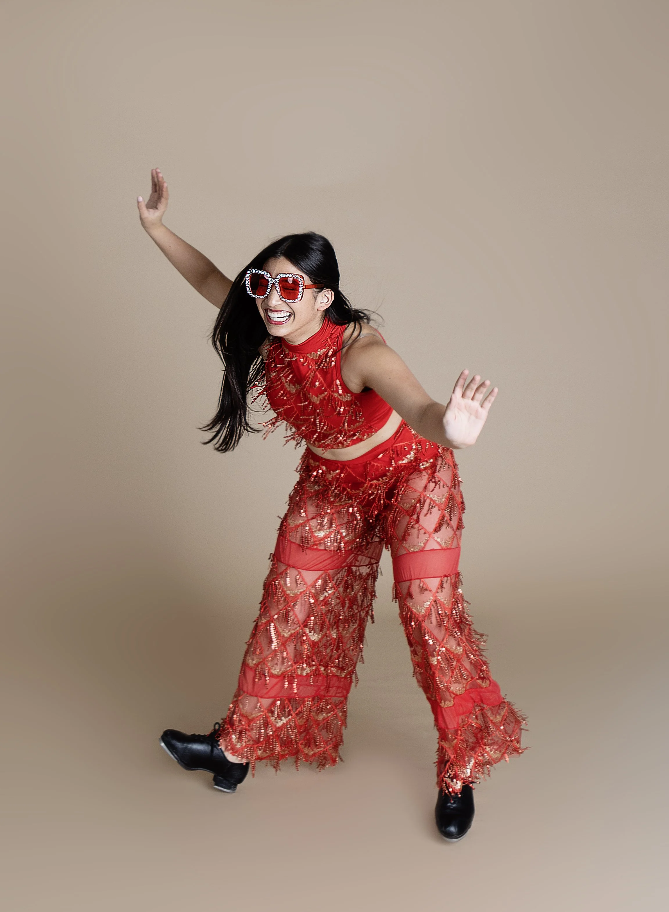 A woman wearing a red, sparkly, and sheer outfit with patterned wide-leg pants, black ice skates, red sunglasses, and smiling playfully against a beige background.
