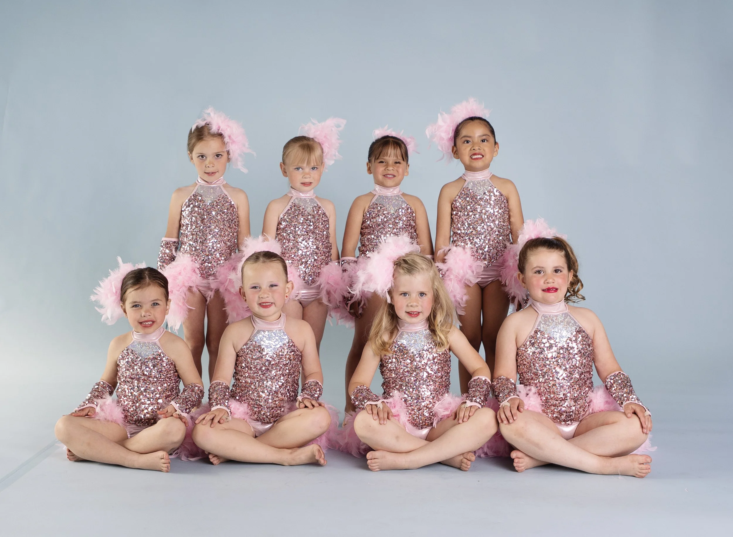 Ten young girls in pink and silver costumes with feathered headpieces, posing for a group photo in a dance studio.