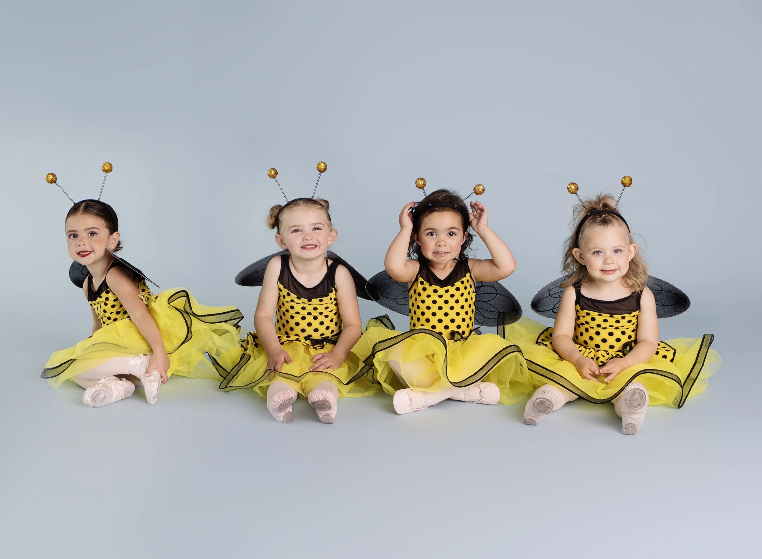 Four young girls dressed as bees in yellow and black costumes with antennae headbands, black wings, and yellow tutus, sitting and posing on a light blue background.