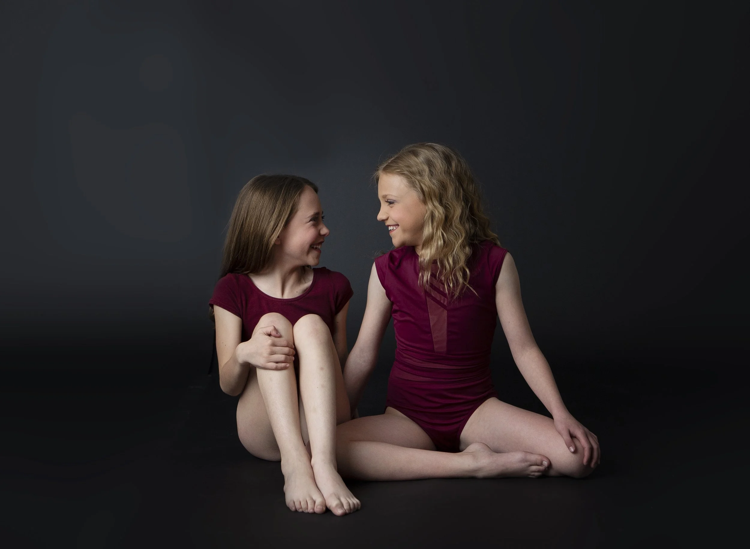 Two young girls sitting on the floor facing each other, smiling and laughing, against a dark background. One girl has straight brown hair and the other has curly blonde hair, both wearing maroon tops.