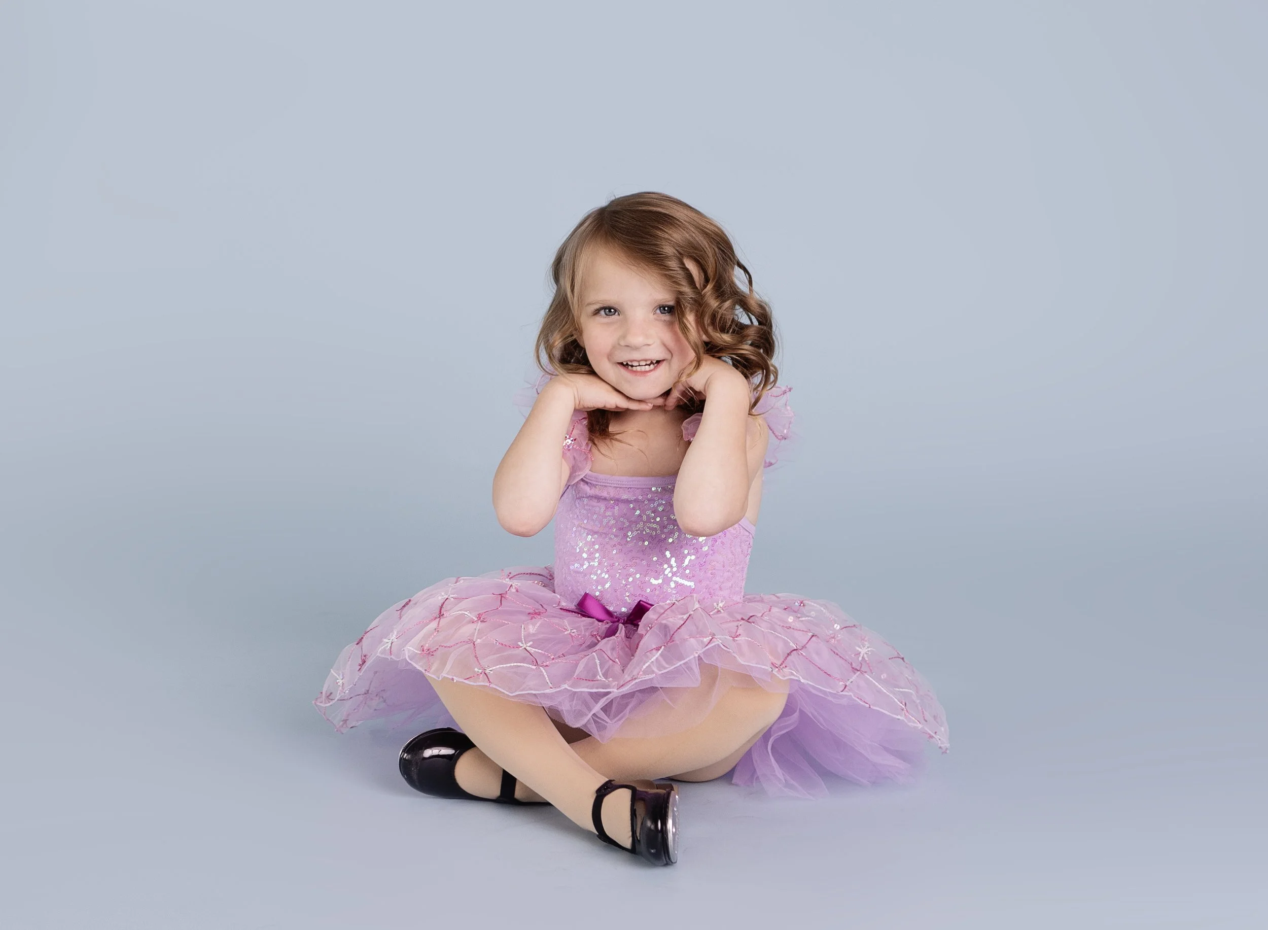 A young girl with curly red hair wearing a pink tutu dress and black shoes, sitting on the floor smiling with her hands near her face against a light blue background.