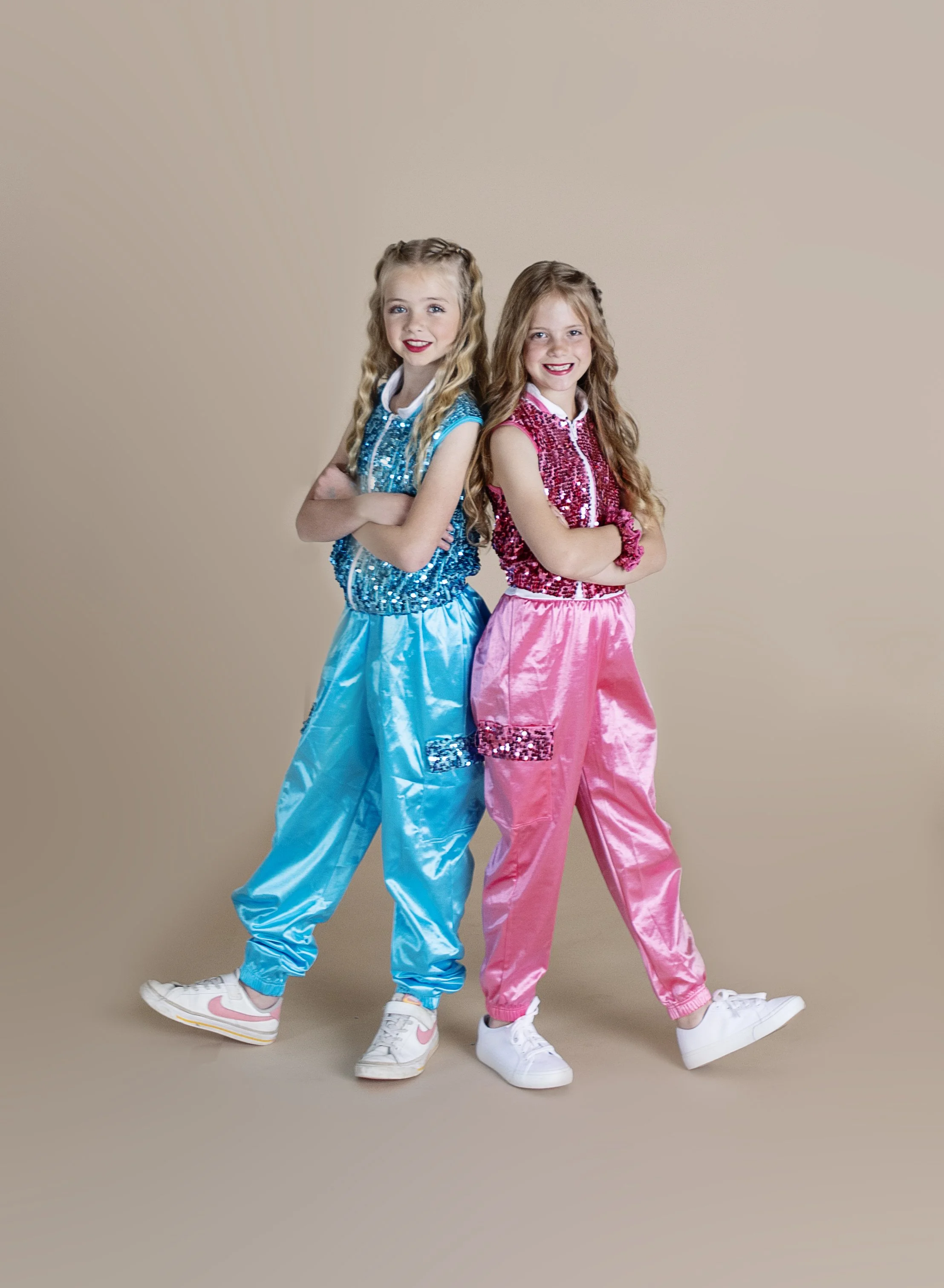 Two young girls with long, wavy hair, crossing arms, wearing colorful shiny dance or performance outfits and sneakers, standing against a neutral background.