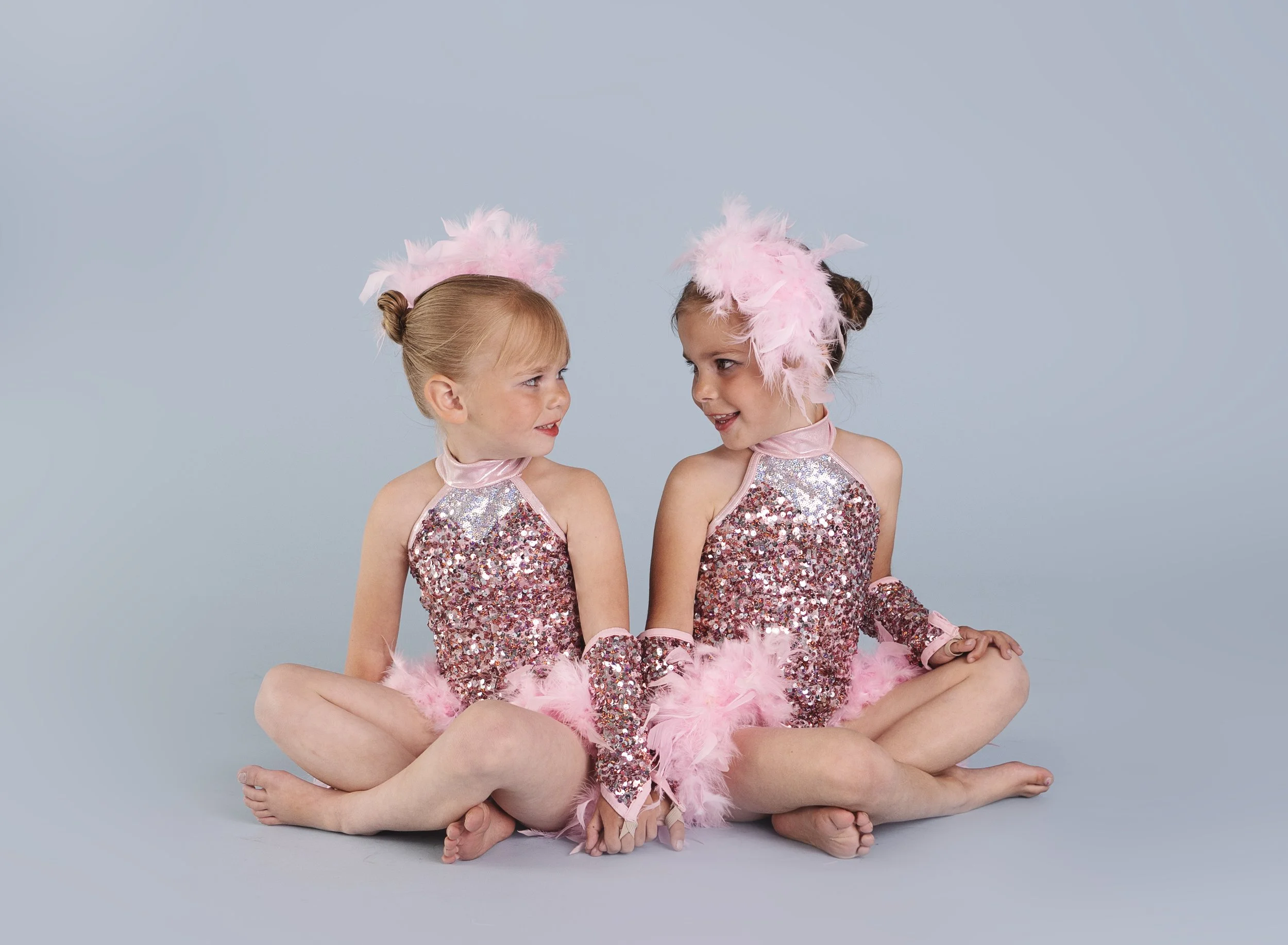 Two young girls in matching pink sequin costumes with feathered headpieces sitting cross-legged and holding hands, engaging in conversation against a plain light blue background.