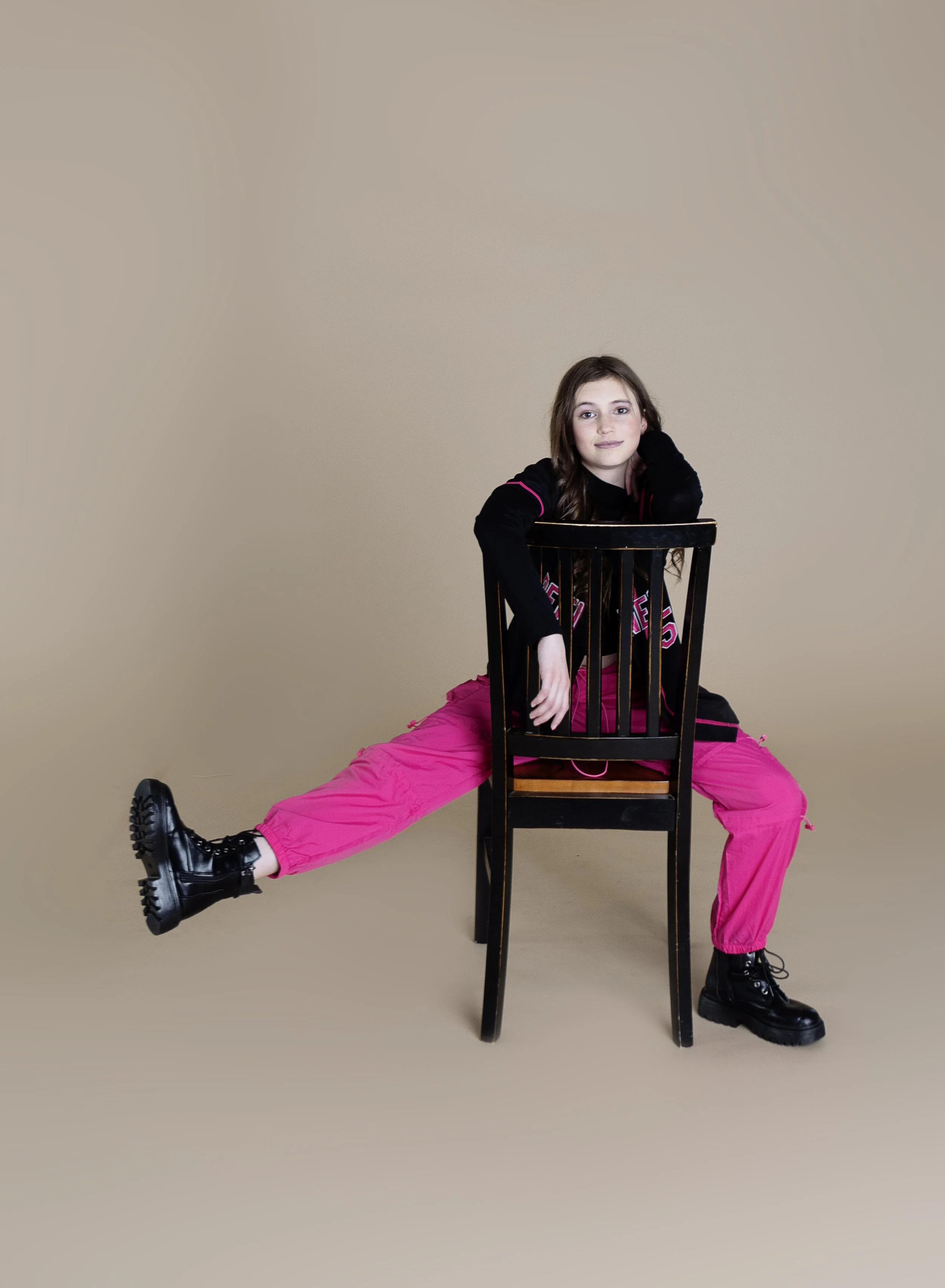 A young woman with long brown hair sitting on a wooden chair, wearing a black jacket with pink accents, pink pants, and black boots. She is posing with one leg extended to the side and resting her head on her hand against the back of the chair.