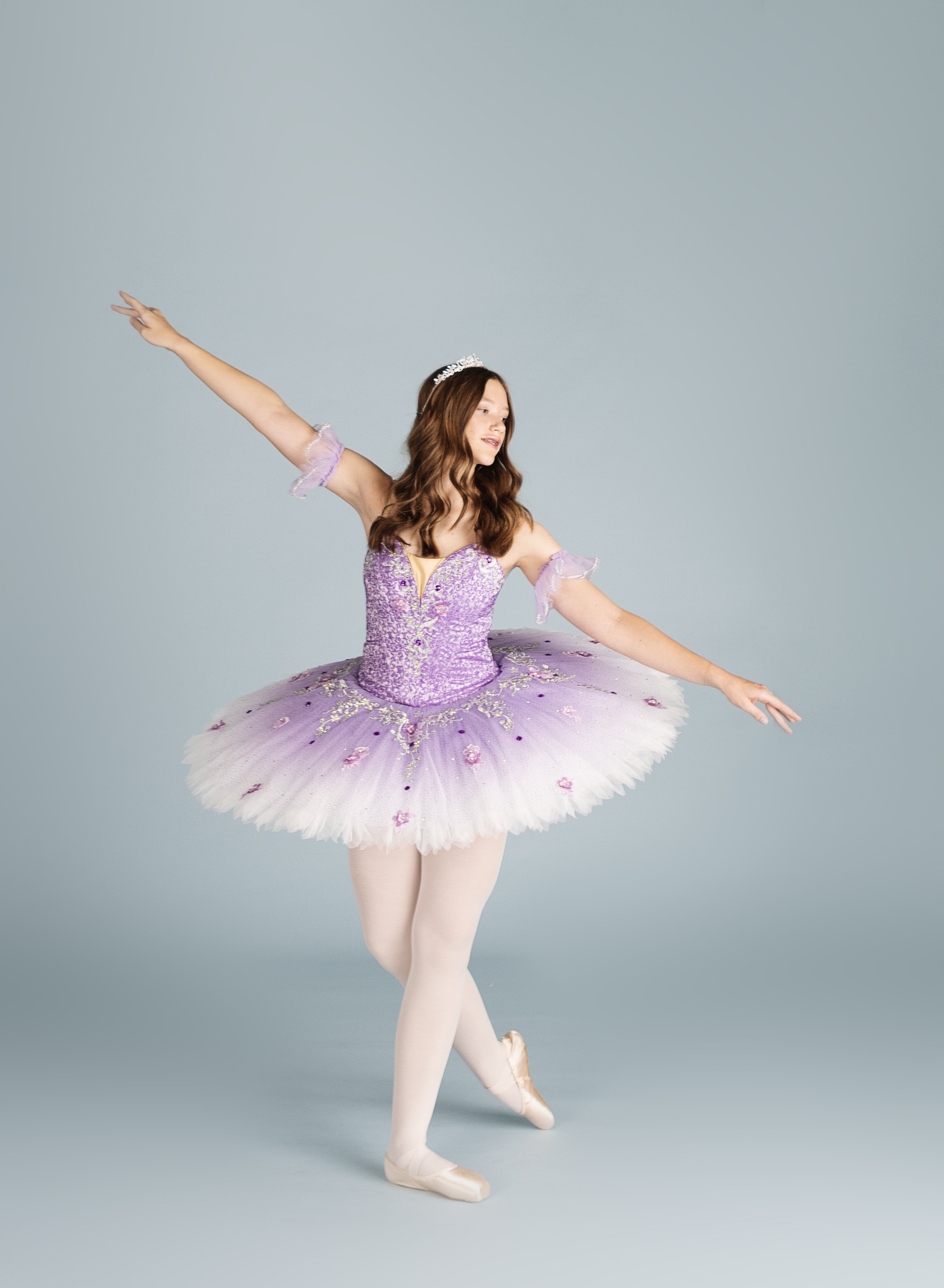 A young woman dressed as a ballerina in a purple tutu with floral embellishments, wearing ballet slippers and a tiara, performing a dance pose against a light gray background.