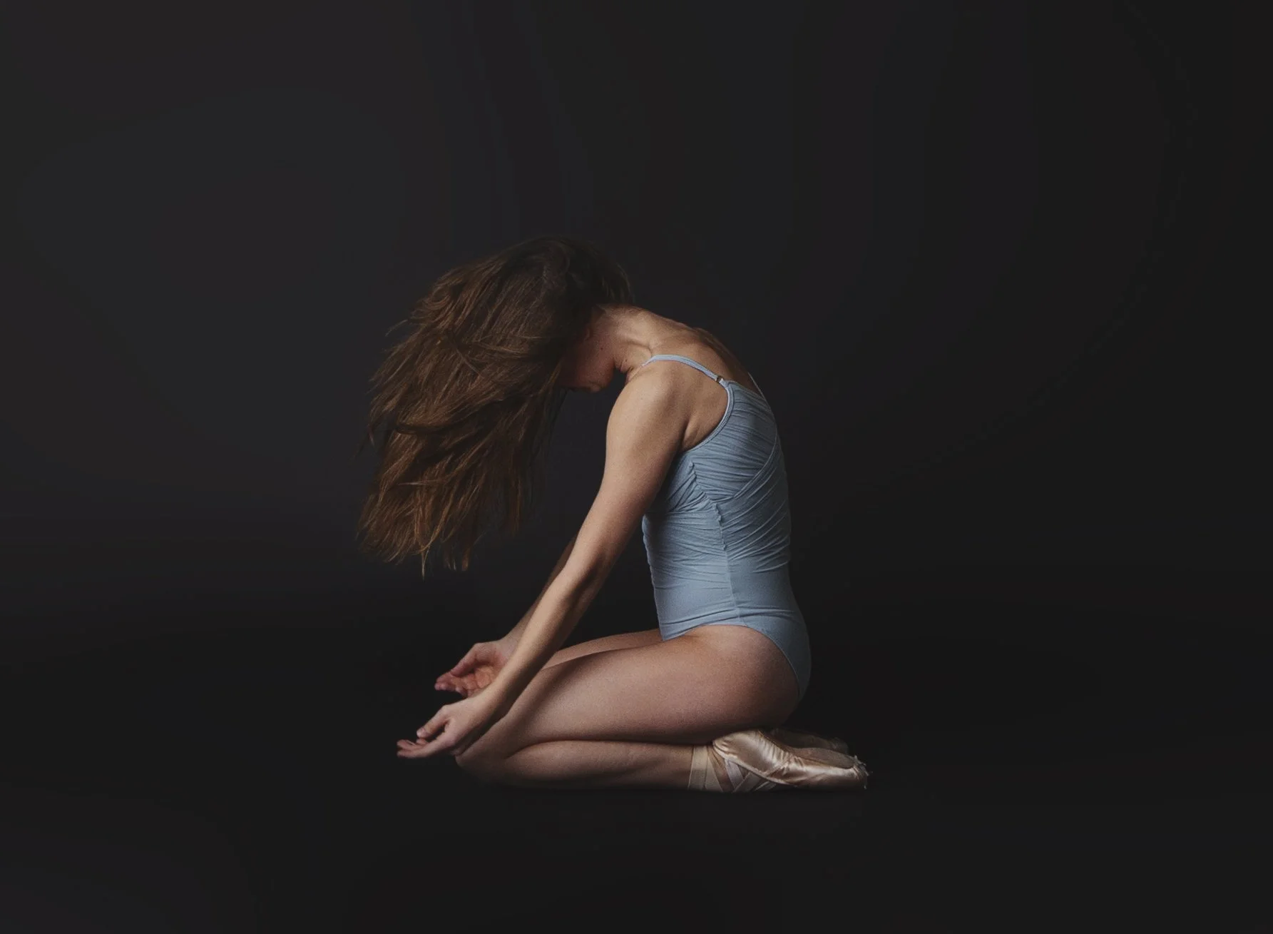 Ballet dancer kneeling with head and hair lowered, wearing a light blue leotard against a dark background.