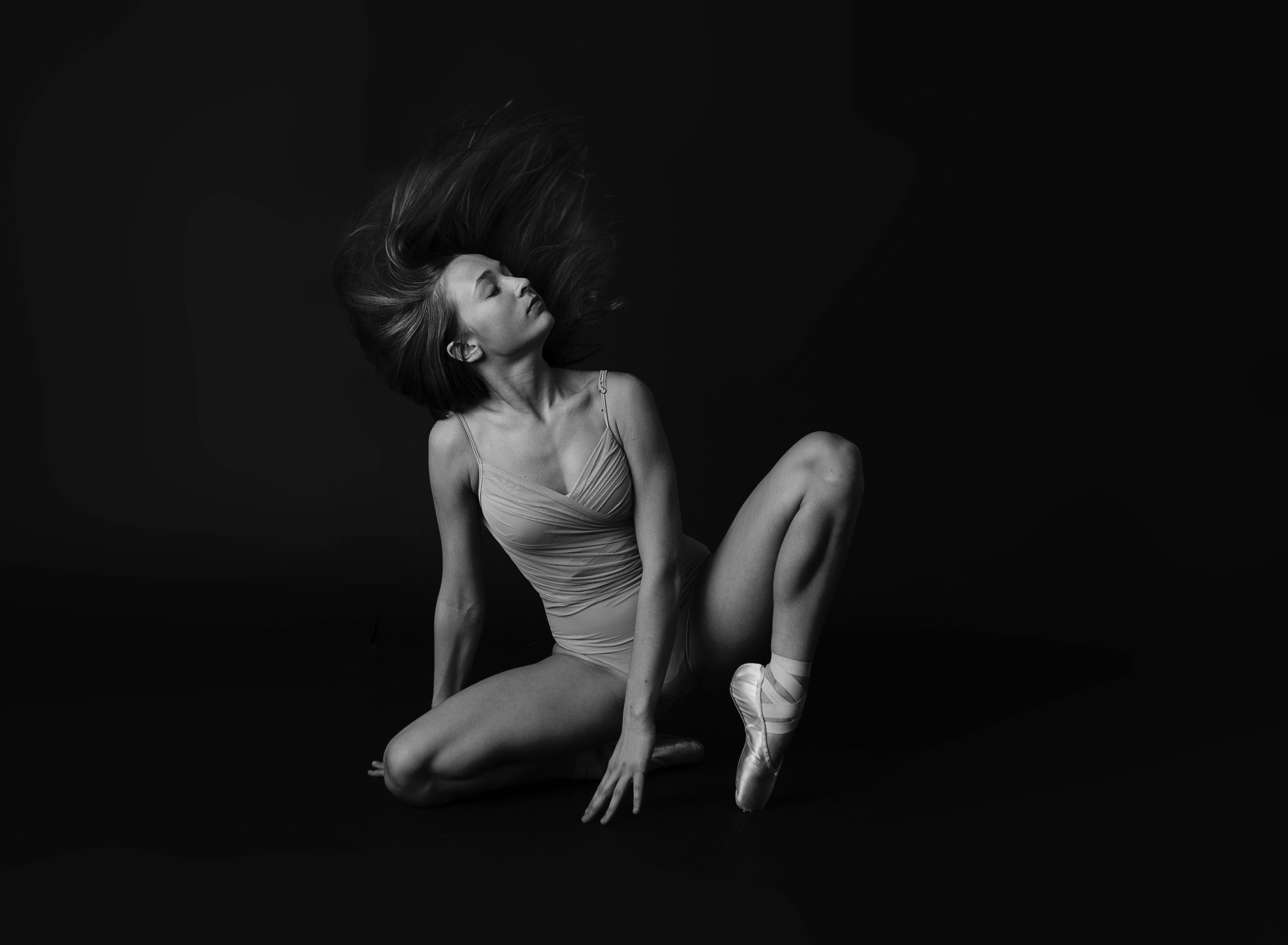 A ballerina with long flowing hair, sitting on the floor, wearing a leotard and pointe shoes, in a black and white photo with a dark background.