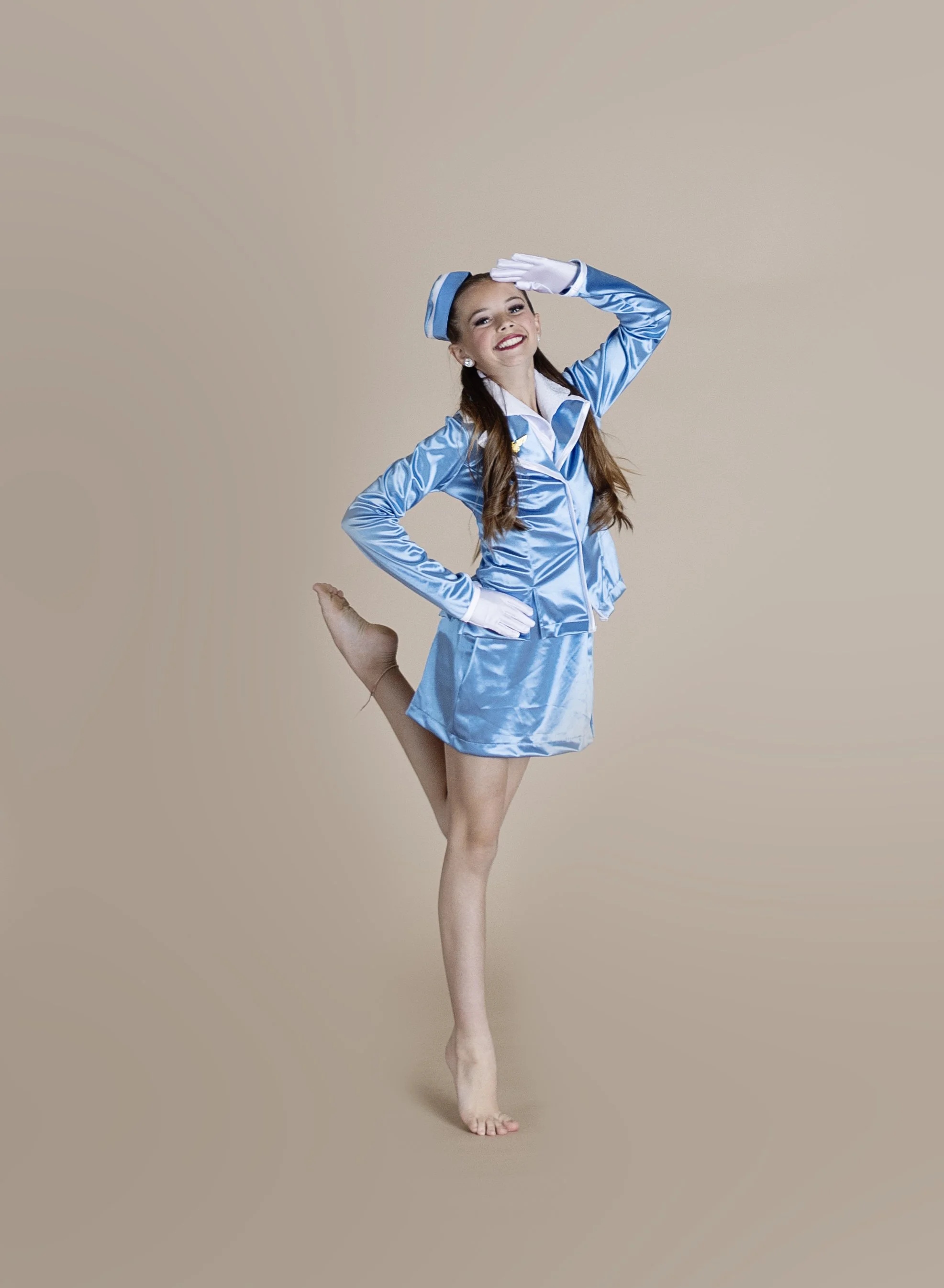A young woman dressed as a flight attendant in a shiny blue uniform, standing on one leg with the other bent behind her, saluting and smiling.