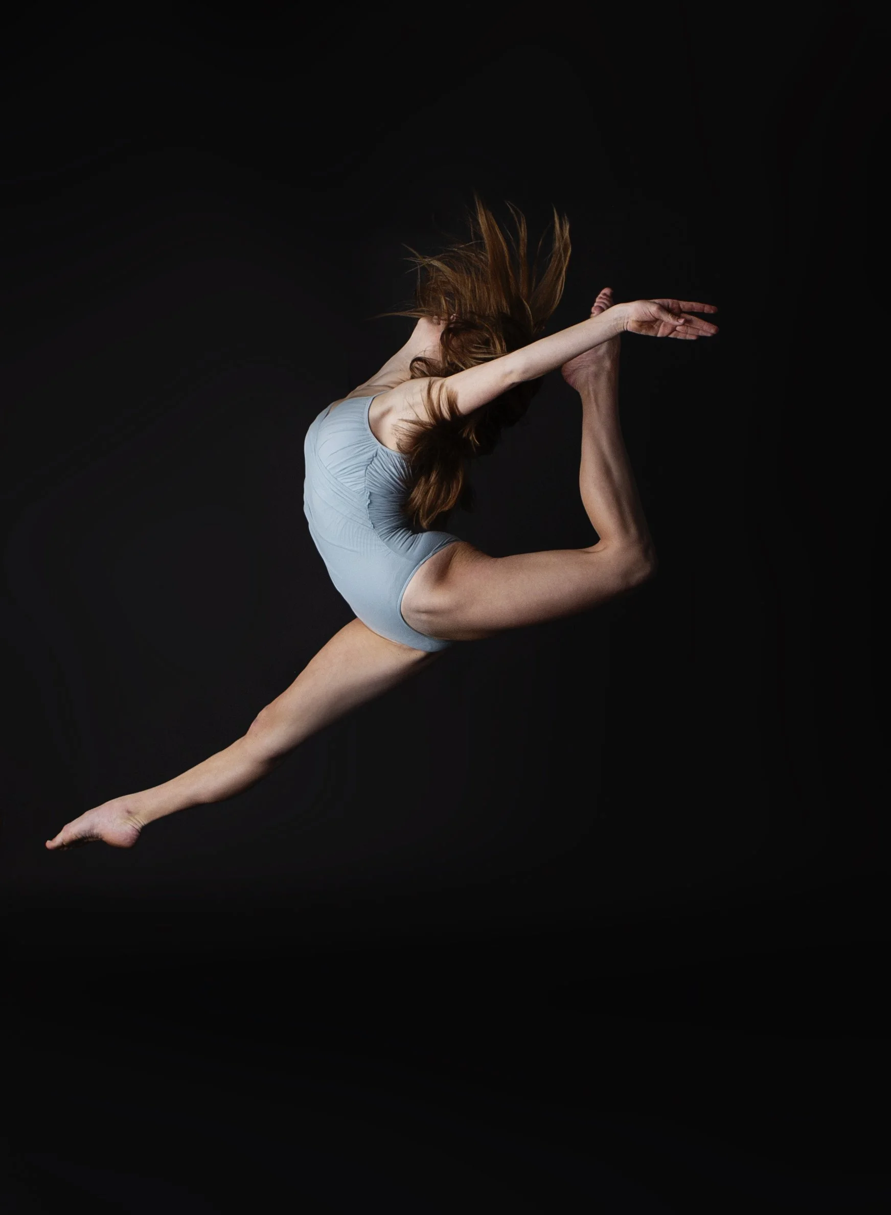 A woman in a light blue leotard performing a complex gymnastics or dance move against a black background, with her body mid-air and her hair flowing.
