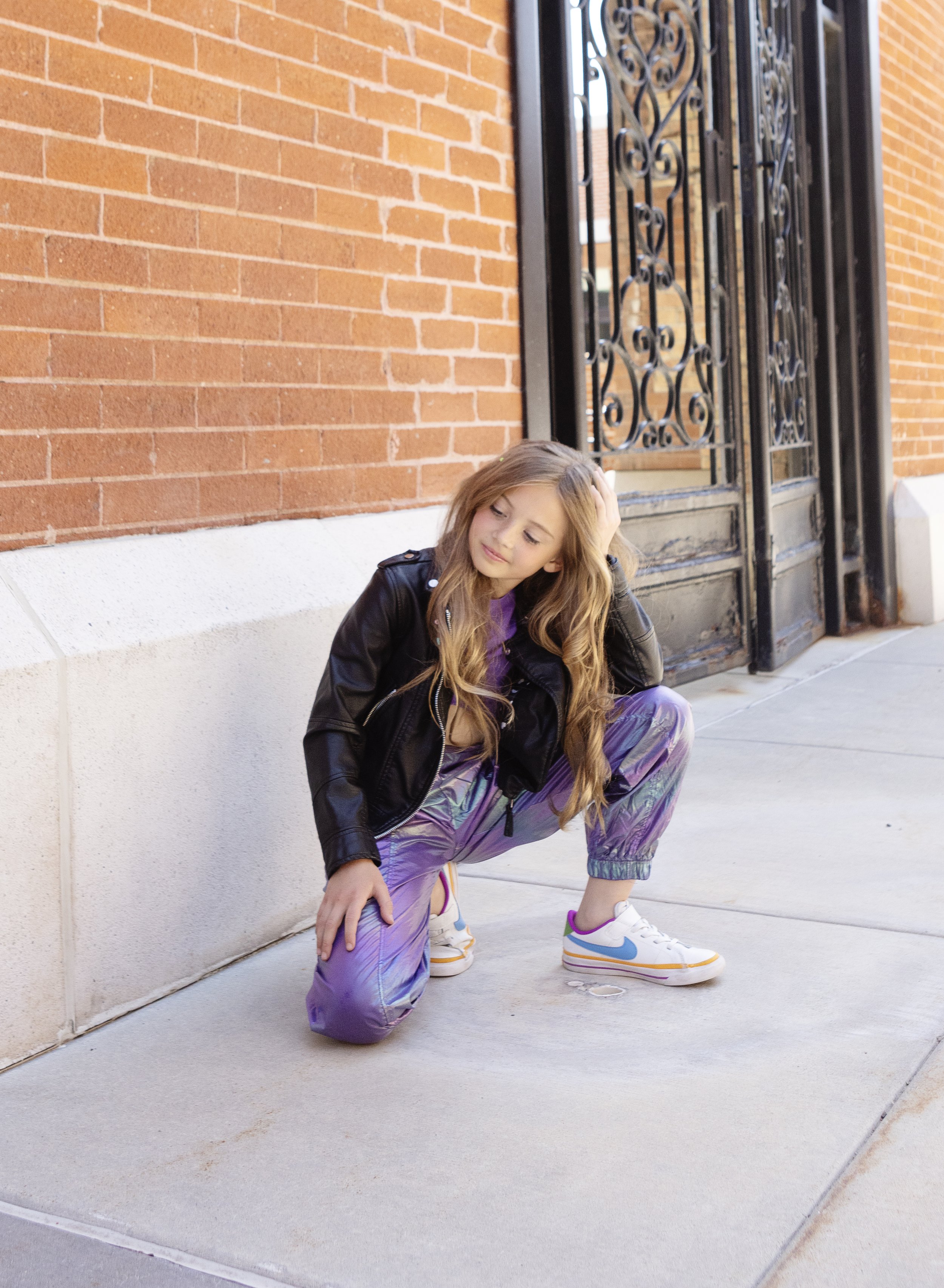Young girl with long wavy hair wearing a black leather jacket, purple shiny pants, and white sneakers squatting on sidewalk near brick wall and black wrought iron gate.