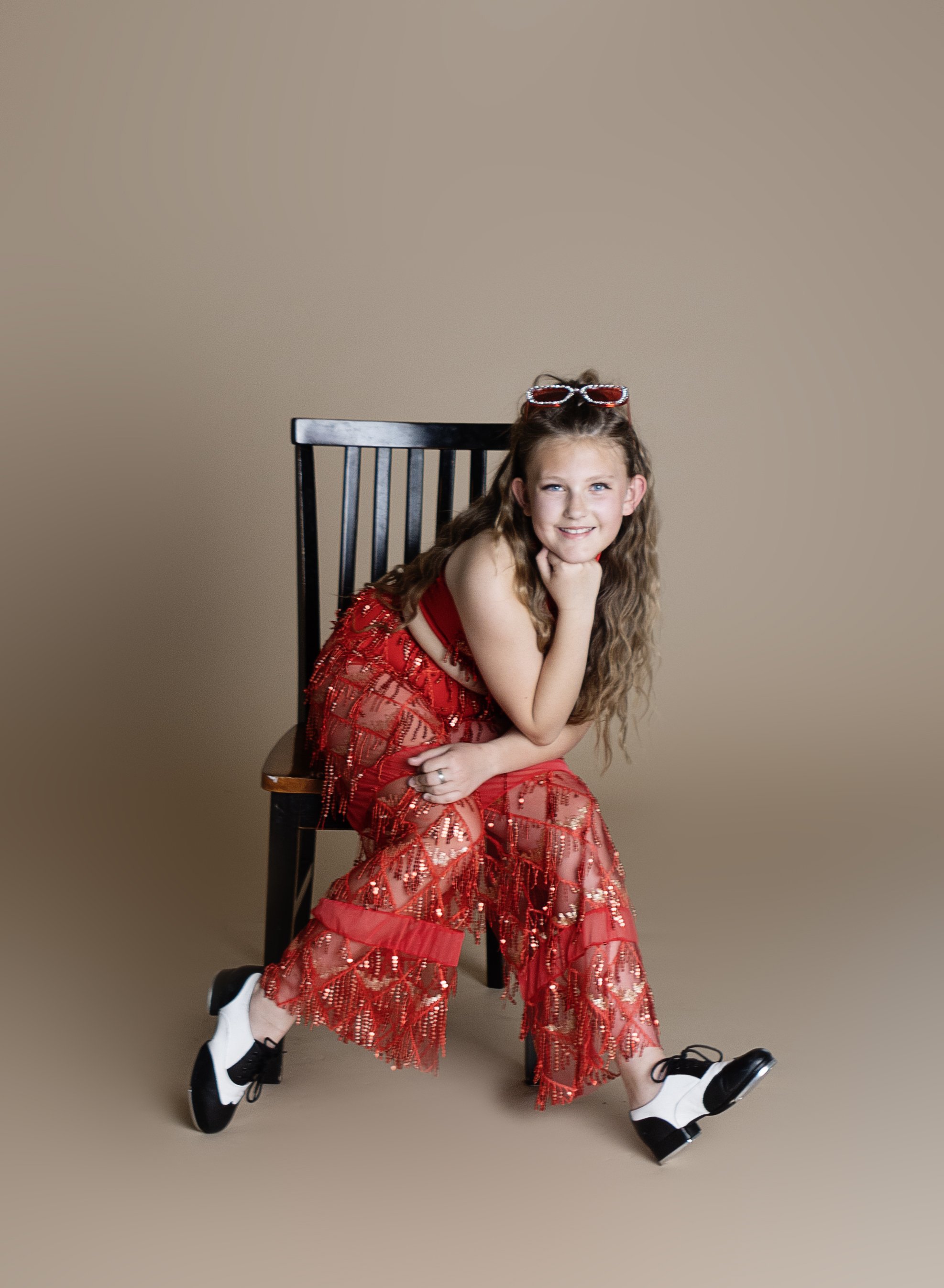 A girl with long curly hair sitting on a black wooden chair, wearing a red semi-transparent sequined dress with pants underneath, white and black dance shoes, and red sunglasses on her head, smiling at the camera against a plain beige background.