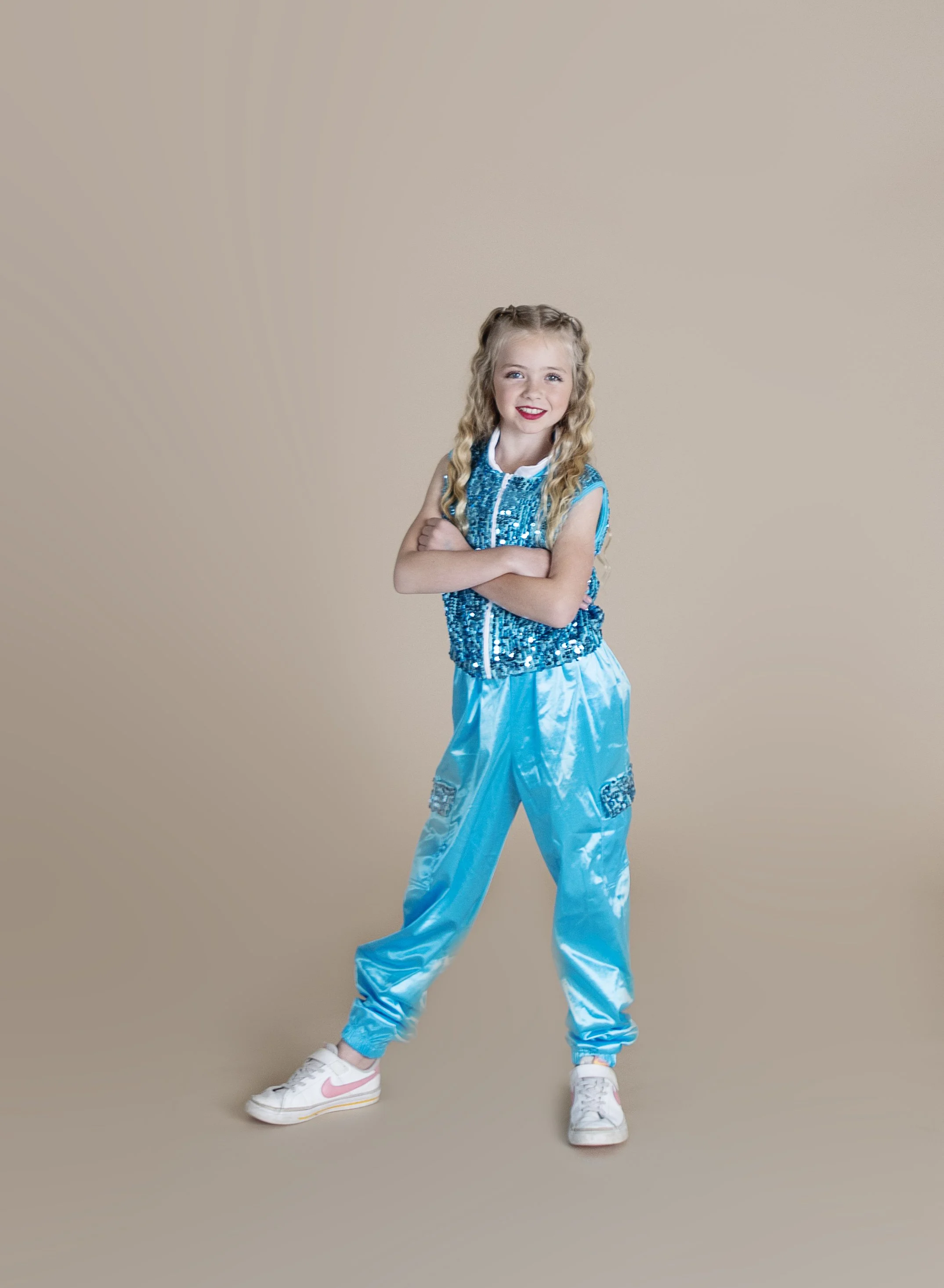A young girl with long curly blonde hair, wearing a blue sequined top and shiny light blue pants, stands with arms crossed and smiles at the camera.