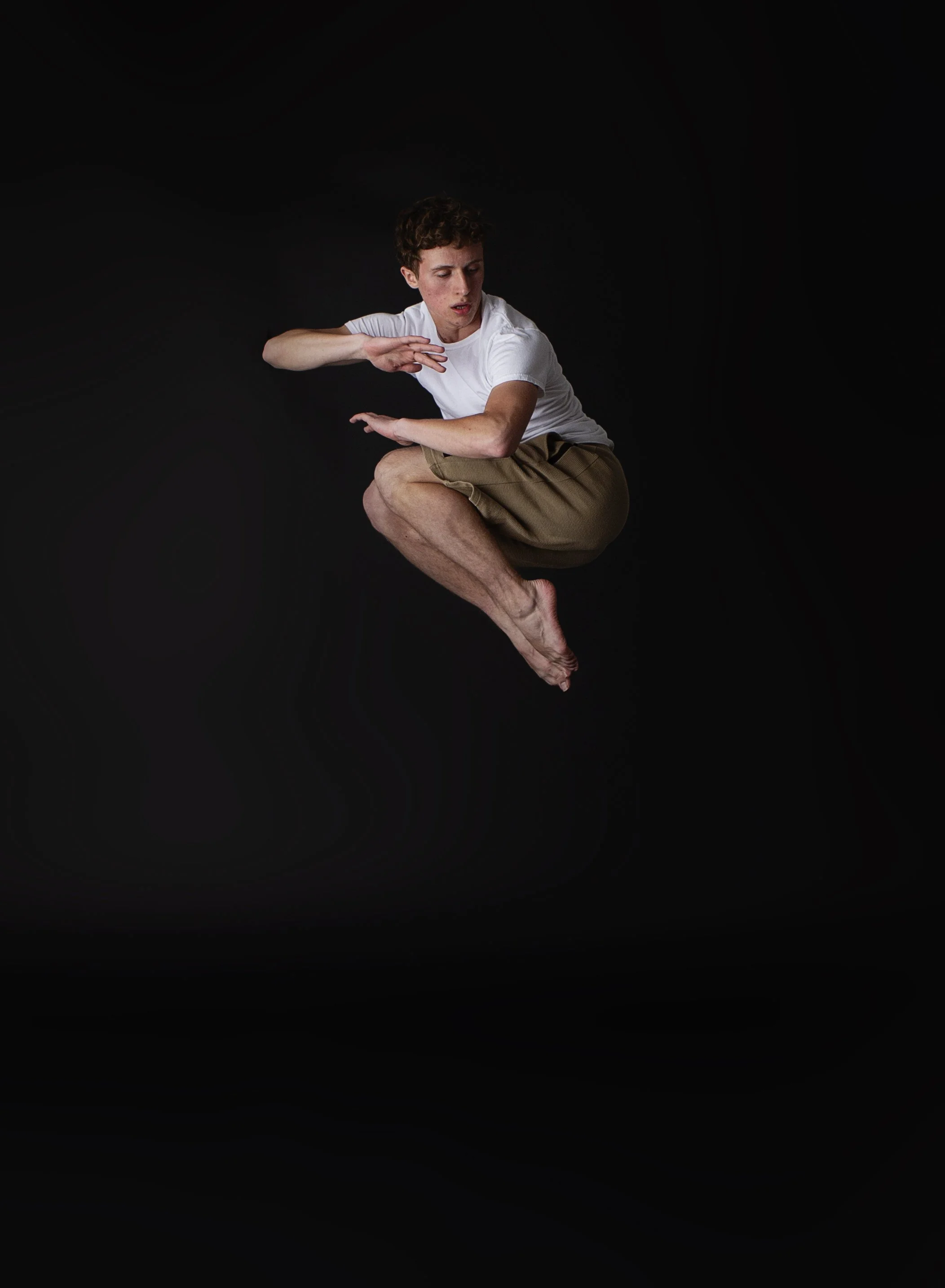 A young man with curly hair, wearing a white t-shirt and khaki shorts, is sitting in a curled position with his eyes closed against a black background.