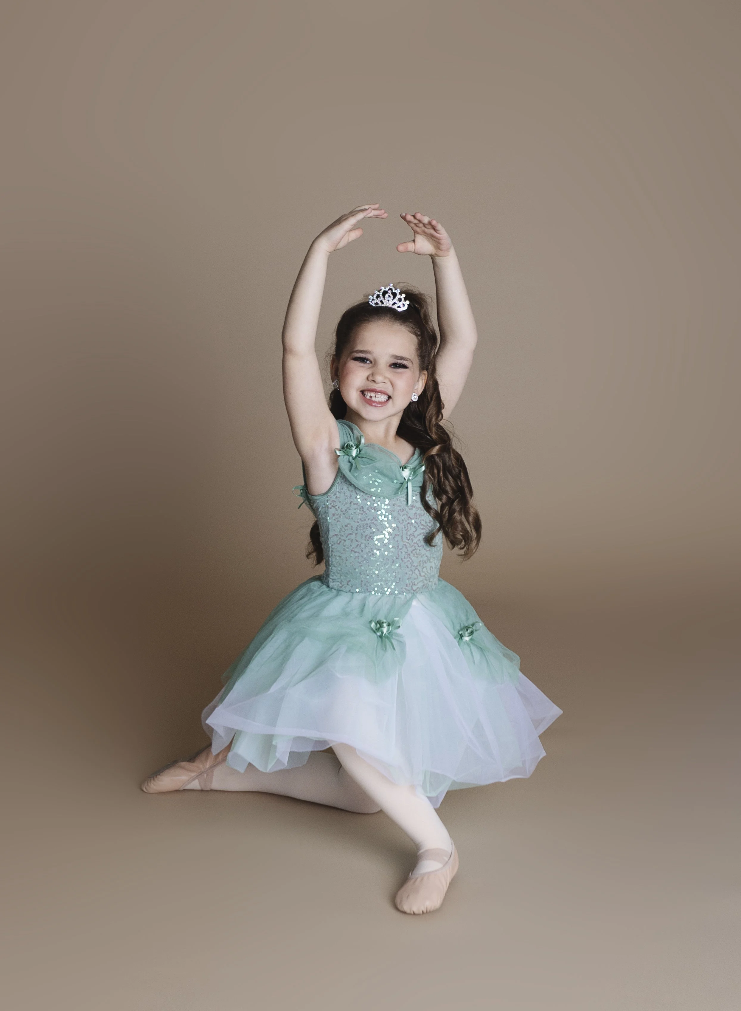 A young girl dressed as a ballerina, wearing a blue tutu and a tiara, is kneeling on the floor with arms raised above her head in a ballet pose, smiling happily.
