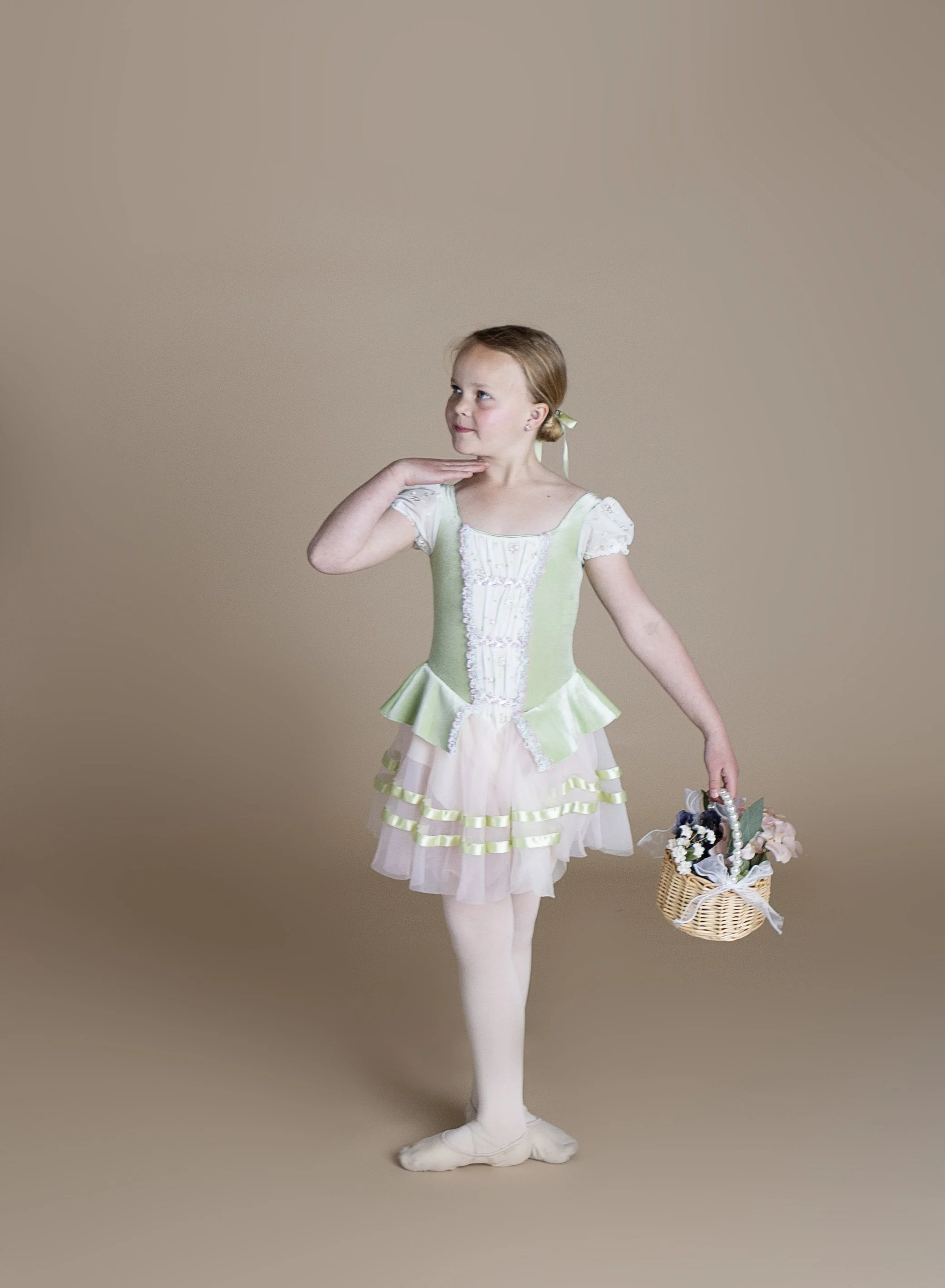 A young girl in a light green and cream dress with ruffles, holding a small basket with flowers, standing on a plain background.