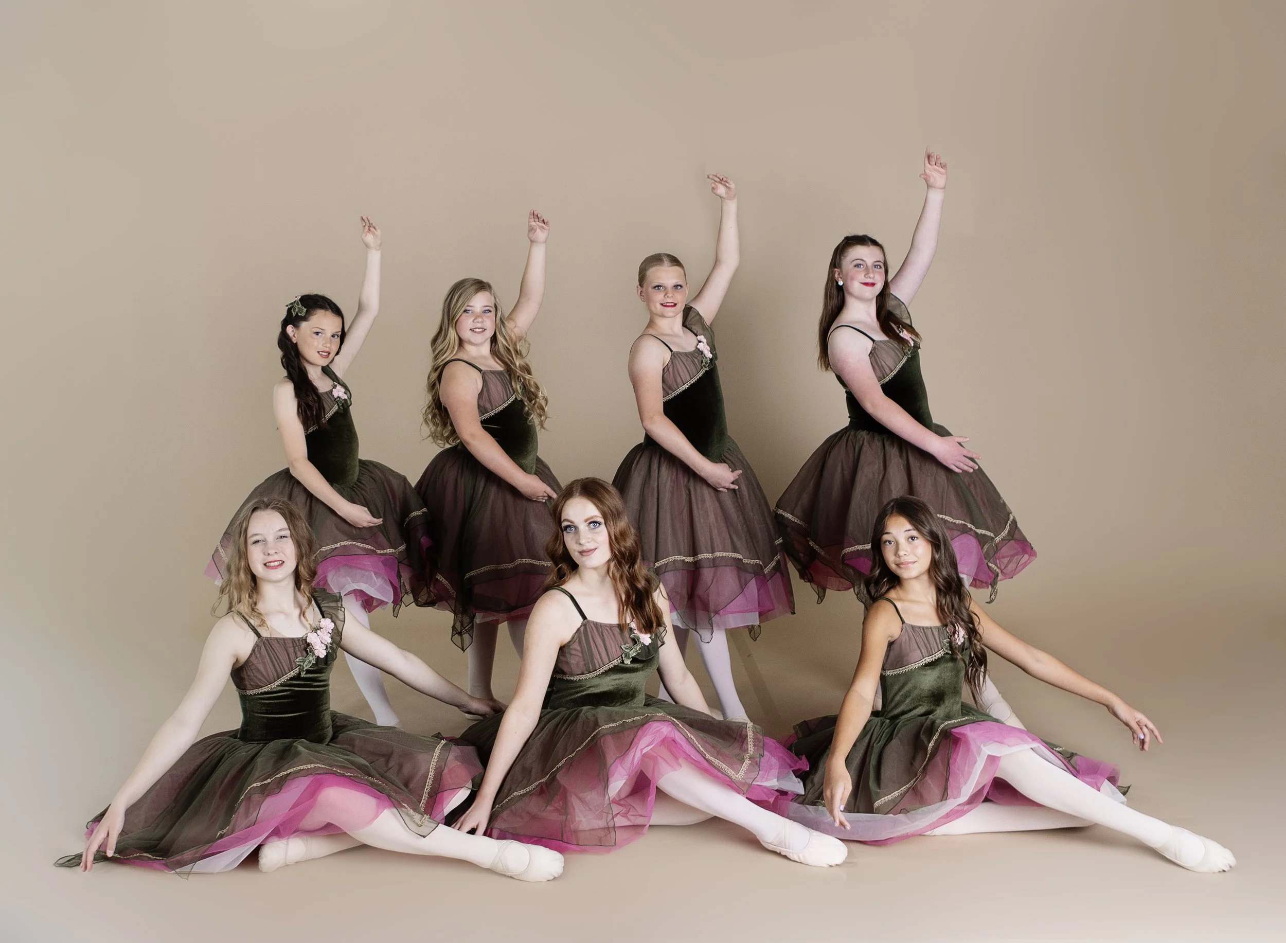 A group of eight young ballet dancers in matching green and pink tutus, posing against a beige background, with some standing and others sitting, all smiling and striking ballet poses.