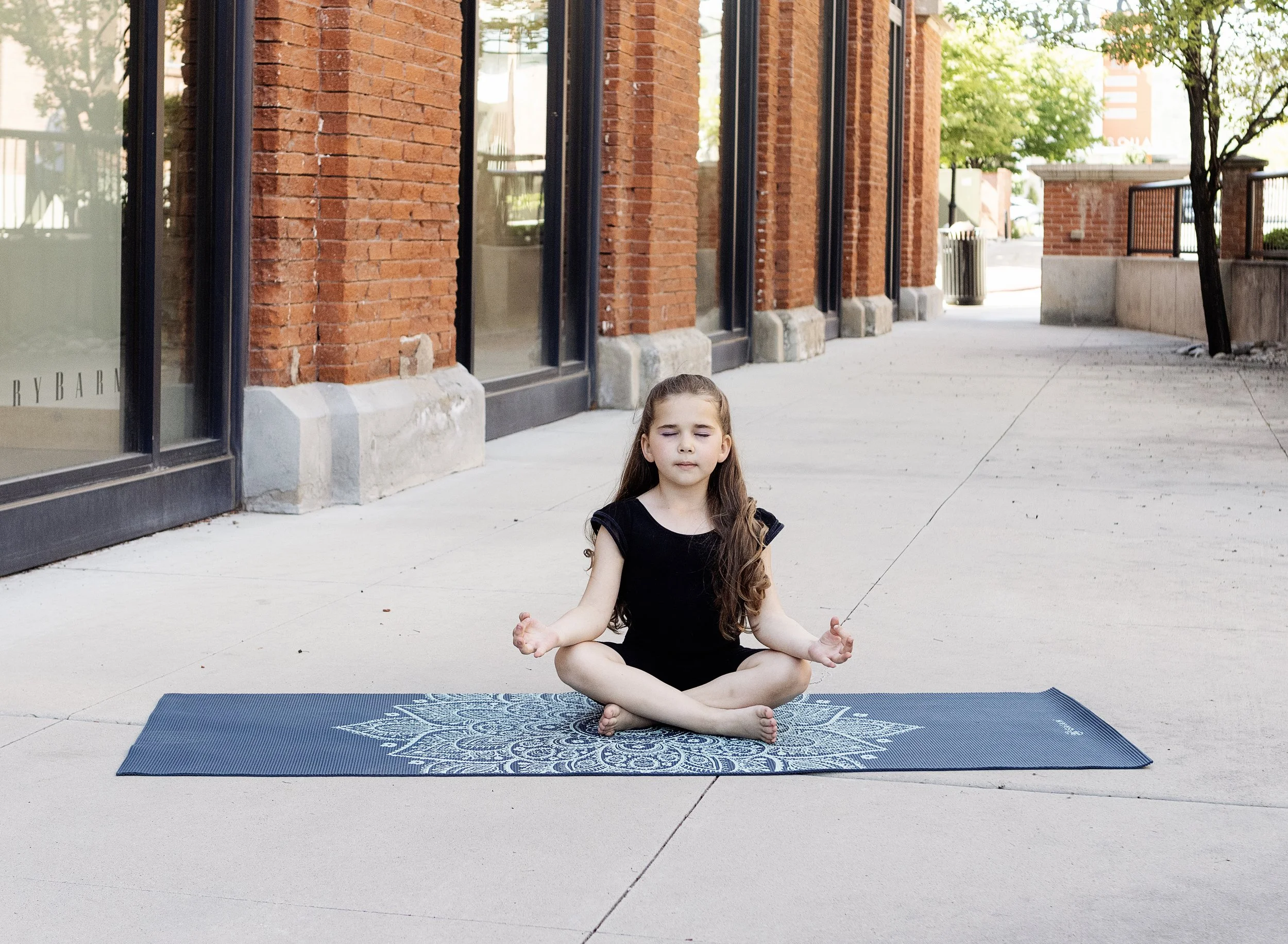 Young girl practicing yoga or meditation on a blue patterned mat outside on a sidewalk, sitting cross-legged with eyes closed, near a brick building with large windows.