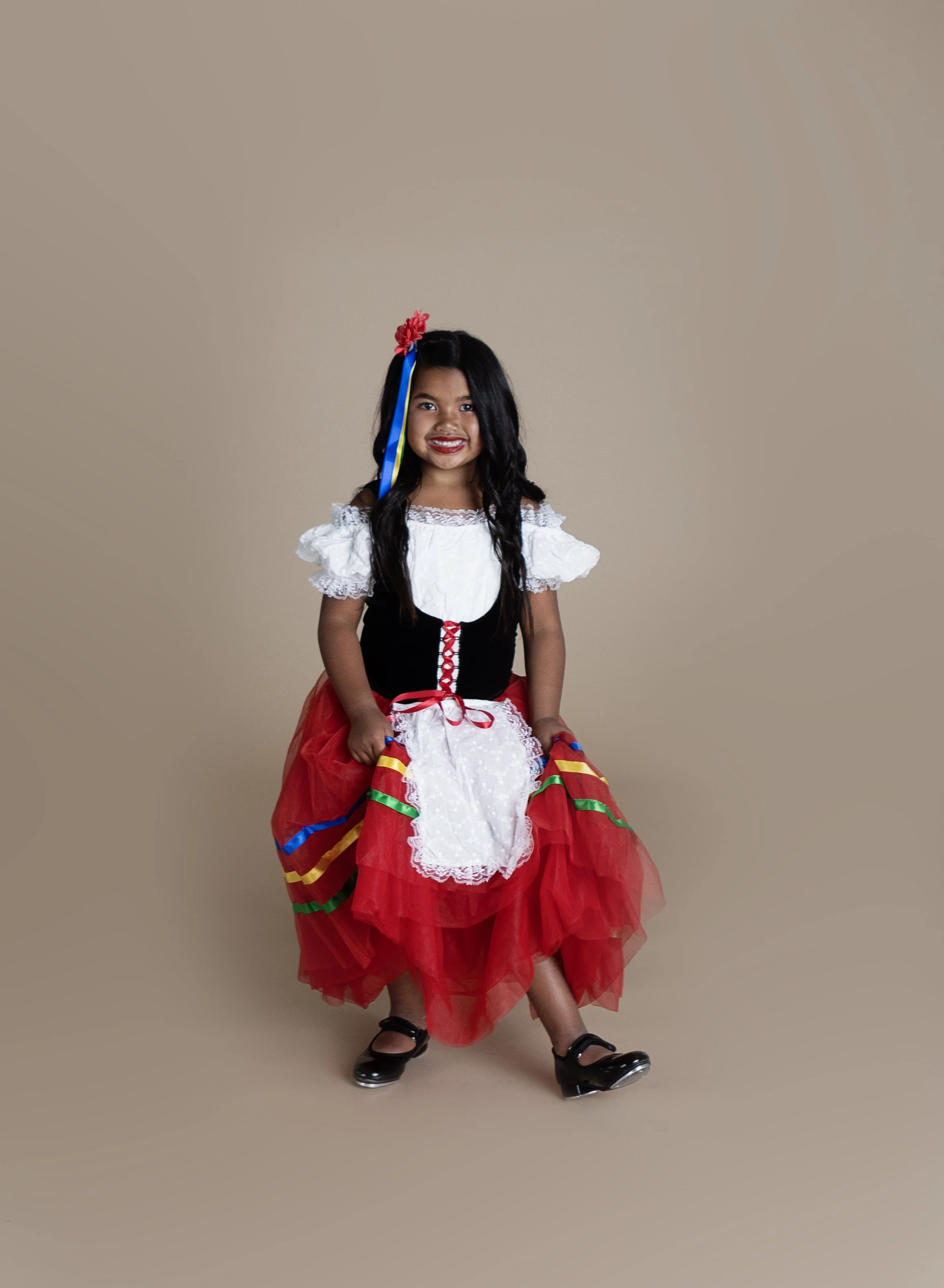 A young girl dressed in traditional Mexican attire, sitting on a stool with a beige background.